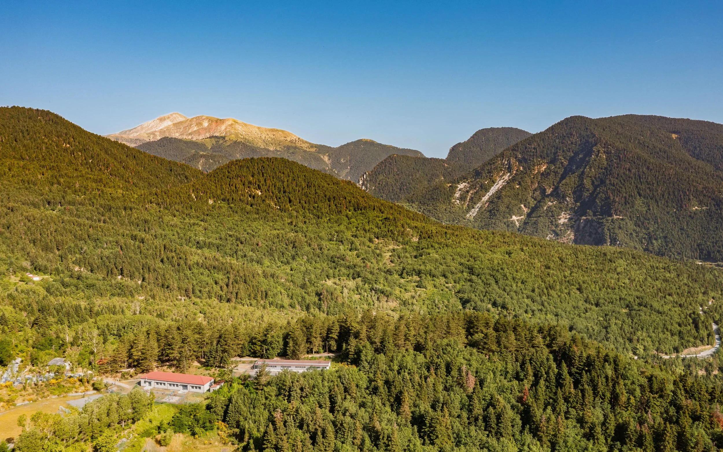 Panoramic view of green forested mountains under a clear blue sky, with a few buildings and a winding road visible in the lower part of the image.