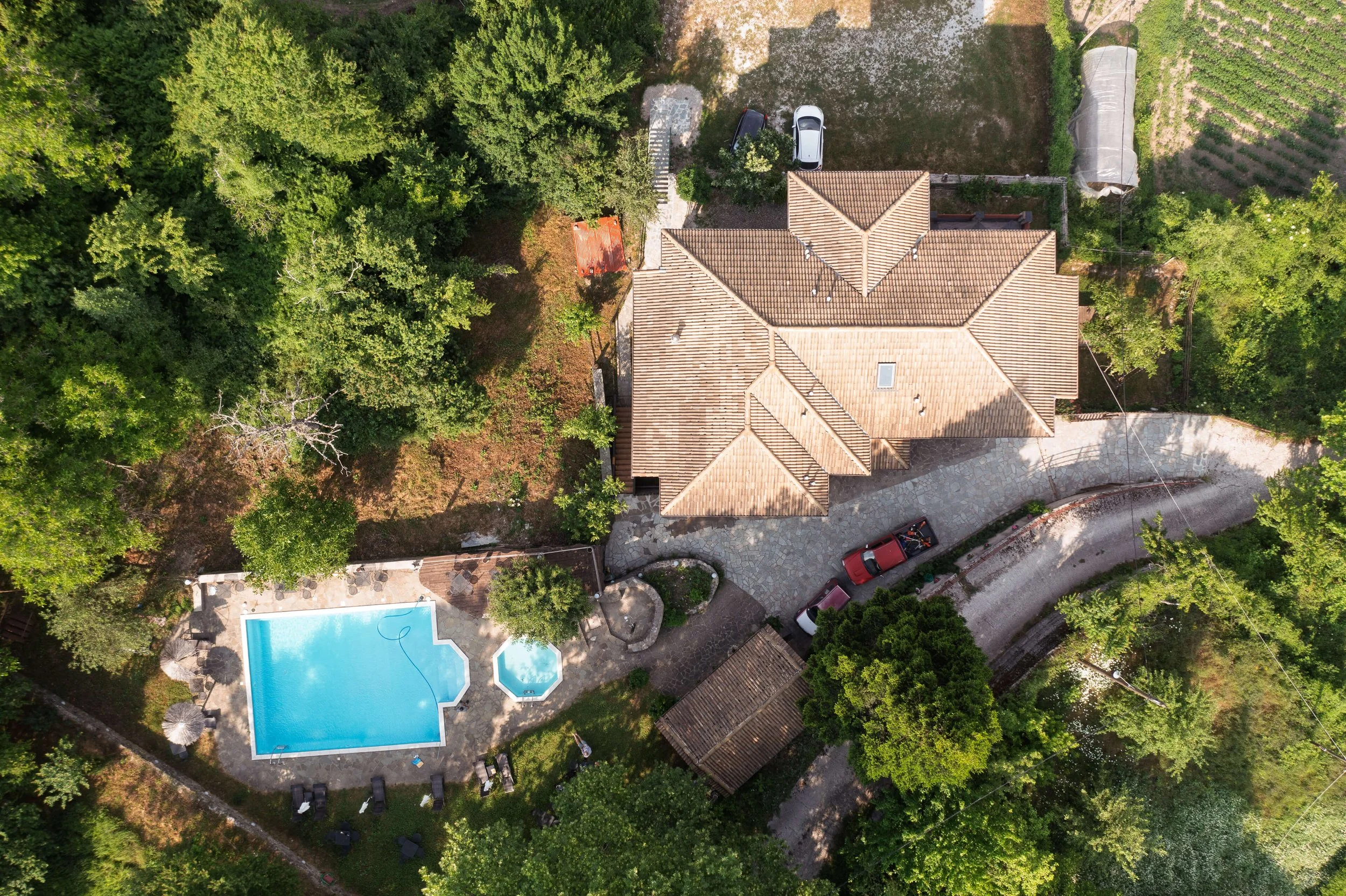 An aerial view of a house with a tiled roof, surrounded by trees, with a driveway leading up to it. There is a swimming pool and a hot tub in the backyard, along with a small patio area.