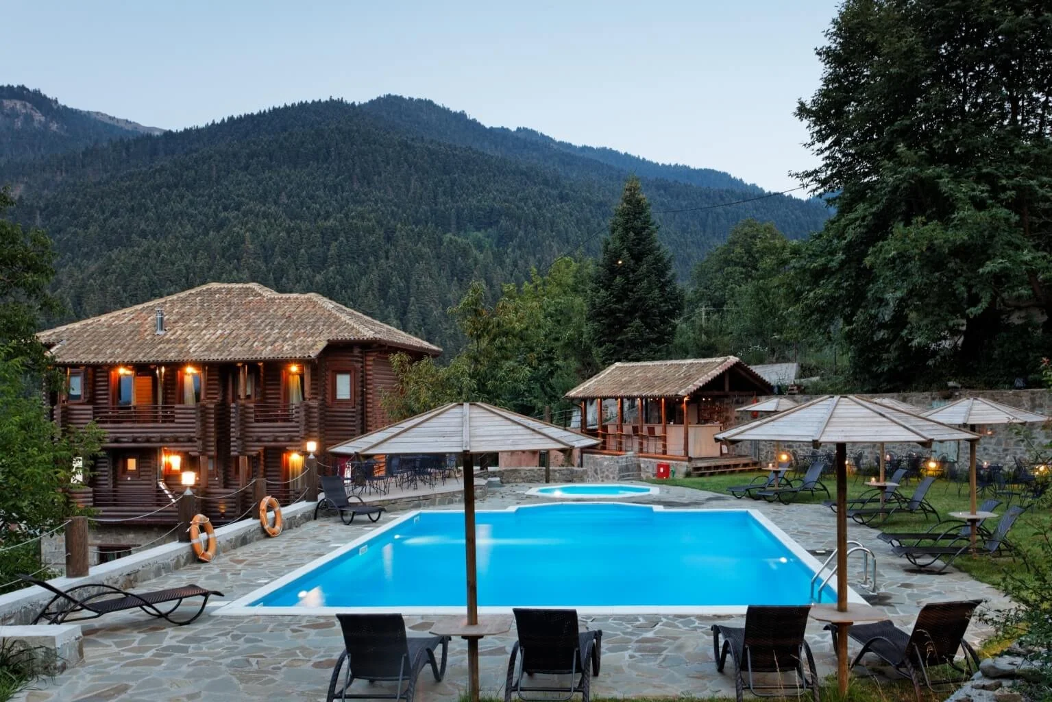 A scenic outdoor pool area at dusk with lounge chairs, umbrellas, and a hot tub, surrounded by a forested mountainous landscape and rustic wooden cabins.