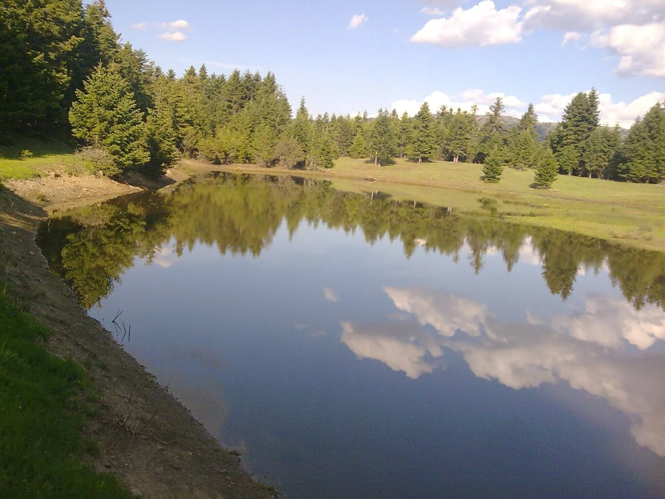 A calm lake with a mirror-like surface reflecting the sky and clouds, surrounded by green grass and trees with a partly cloudy sky overhead.