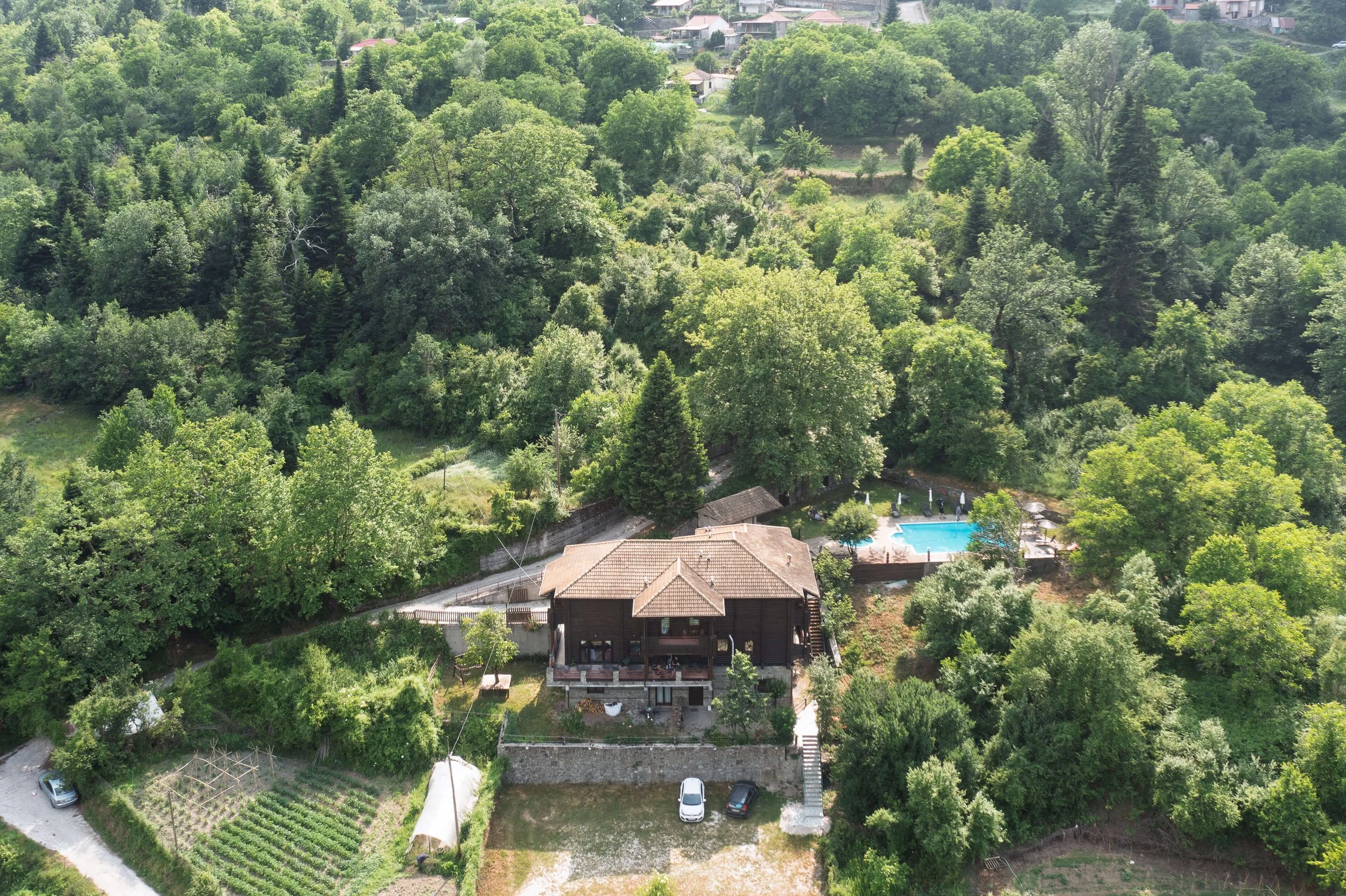Aerial view of a house surrounded by trees, with a swimming pool, a garden with plants, and parked cars visible near the house.