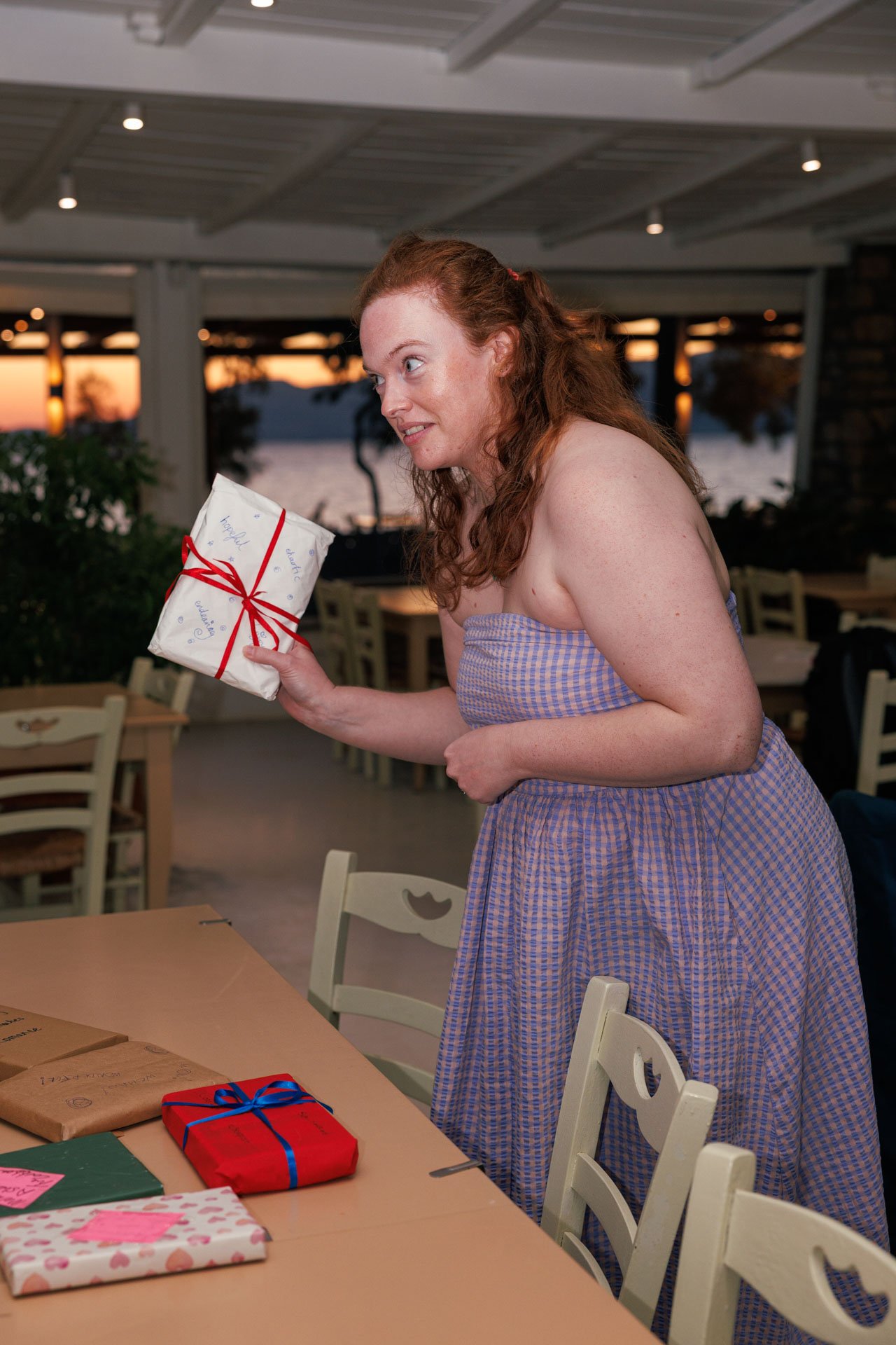A woman with red hair, wearing a purple checkered strapless dress, is holding a small wrapped gift with a red ribbon. She is standing at a table with other wrapped presents, in a restaurant or banquet setting during sunset with a view of water and tr