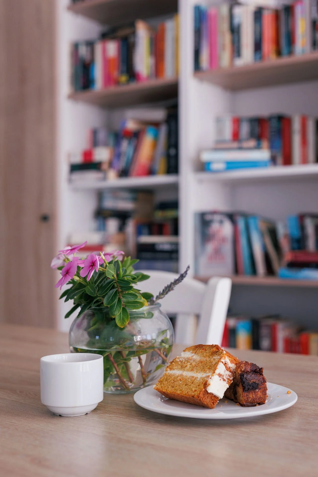 A plate with sliced cake and brownie, a white cup, and a glass jar with a pink flowering plant on a wooden table in a room with bookshelves.