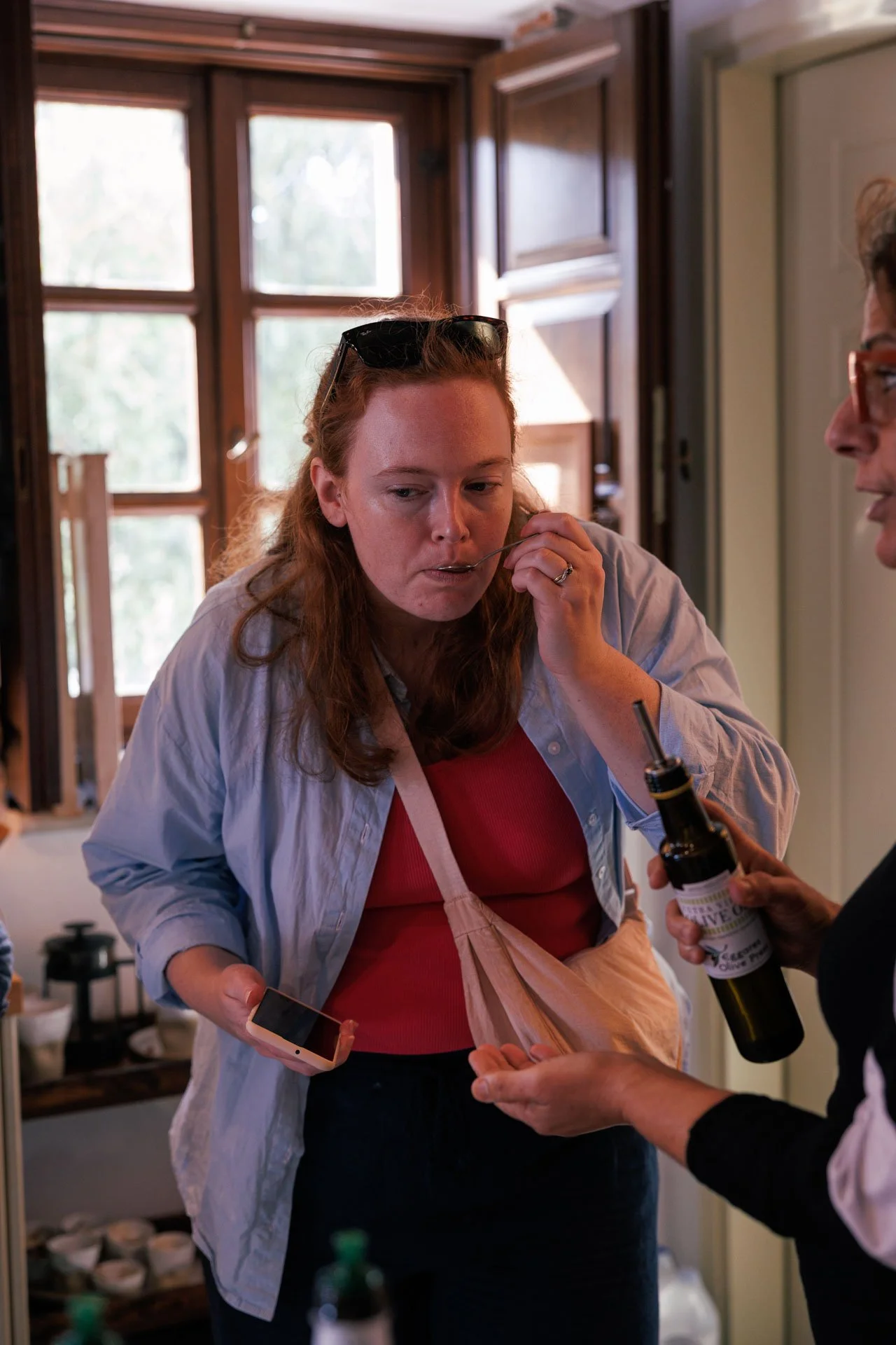 A woman with red hair, wearing a light blue shirt and sunglasses on her head, drinks from a spoon held by another person. The setting appears to be a kitchen with large windows in the background.