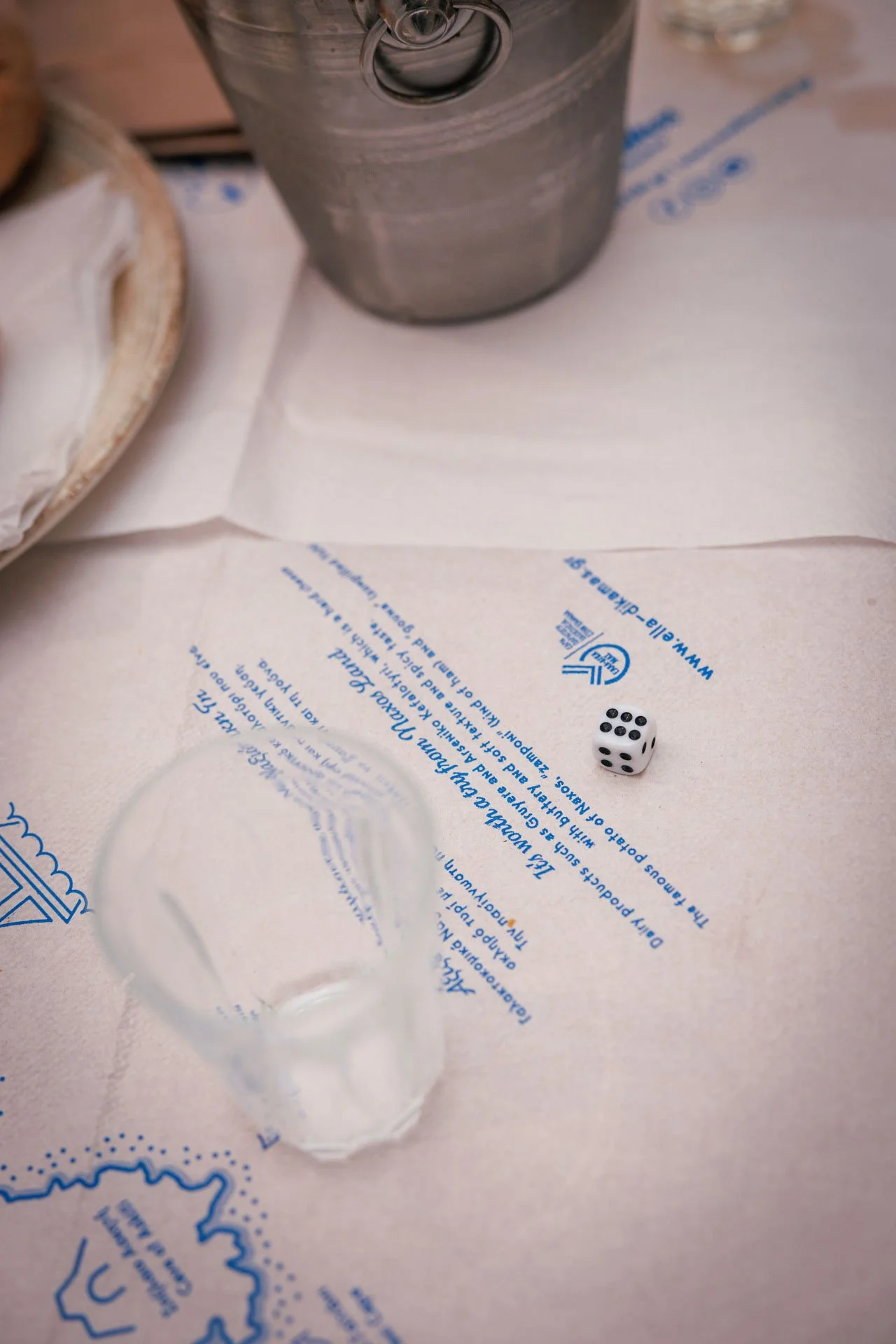 A white paper tablecloth with blue text and a map design, a small clear glass, a white die showing four, and a metal bucket in the background.