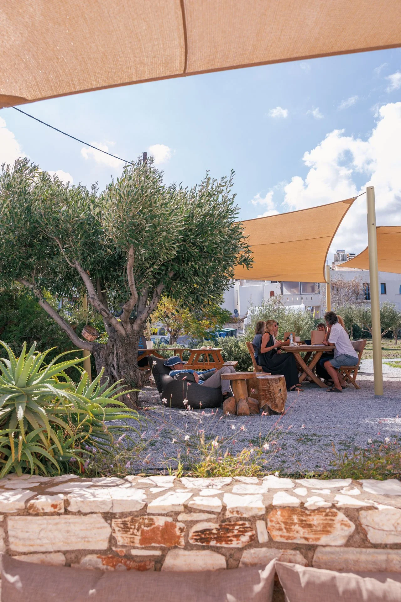 People dining outdoors under tan shade sails, sitting at wooden tables and chairs, with a large tree, green plants, and a stone wall in the foreground, bright blue sky with clouds in the background.