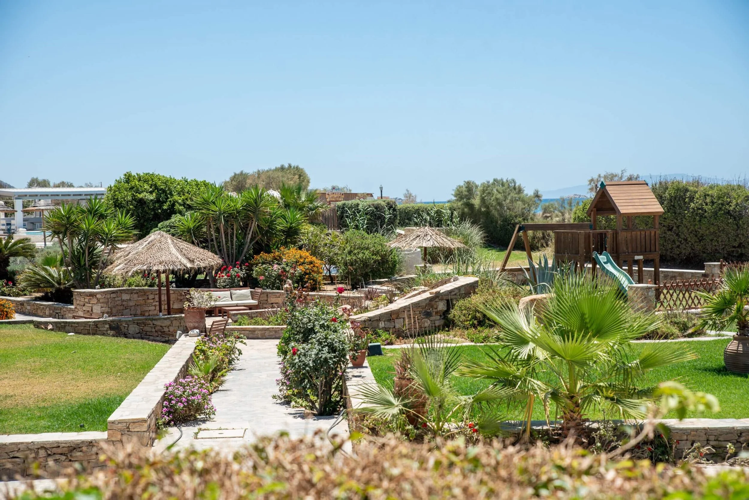 A landscaped garden with palm trees, colorful flowers, a stone pathway, wooden benches with straw umbrellas, and a children's playhouse with slides under a clear blue sky.