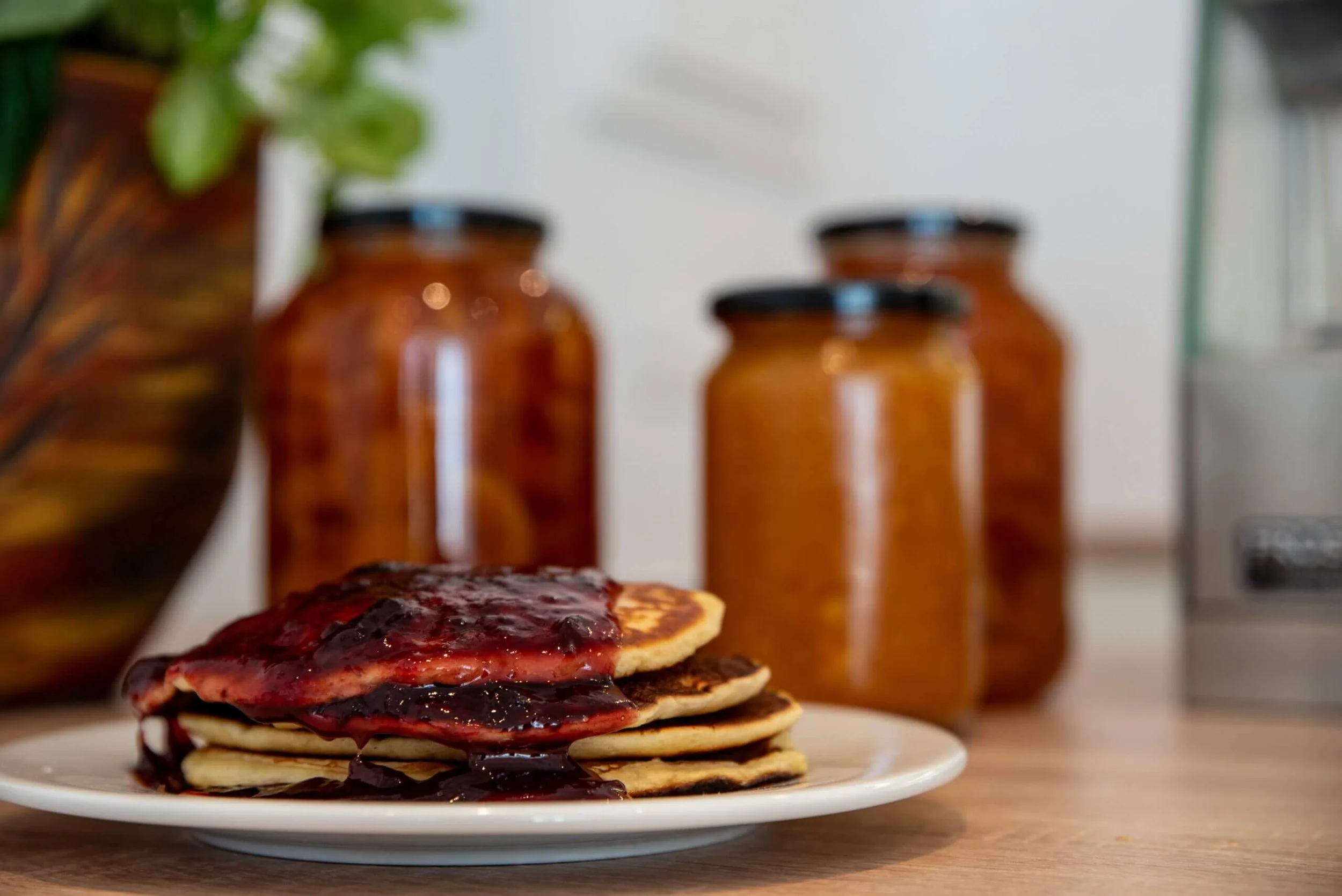 Stack of pancakes topped with blueberry syrup on a white plate, with jars of preserves and a vase with leaves in the background.