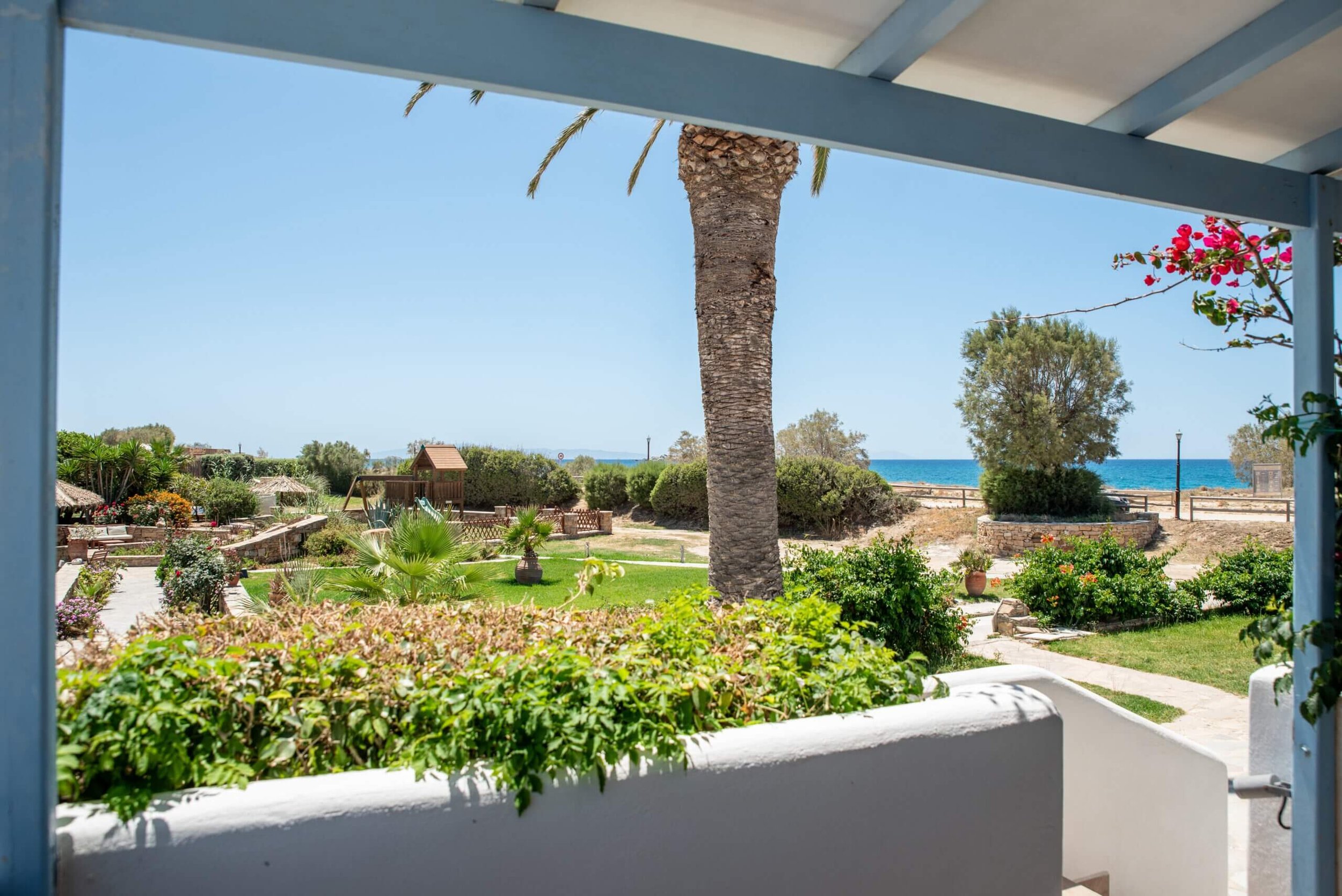 View of a lush garden with a large palm tree, colorful flowers, bushes, a wooden playhouse, and the ocean in the background, framed by a shaded veranda.