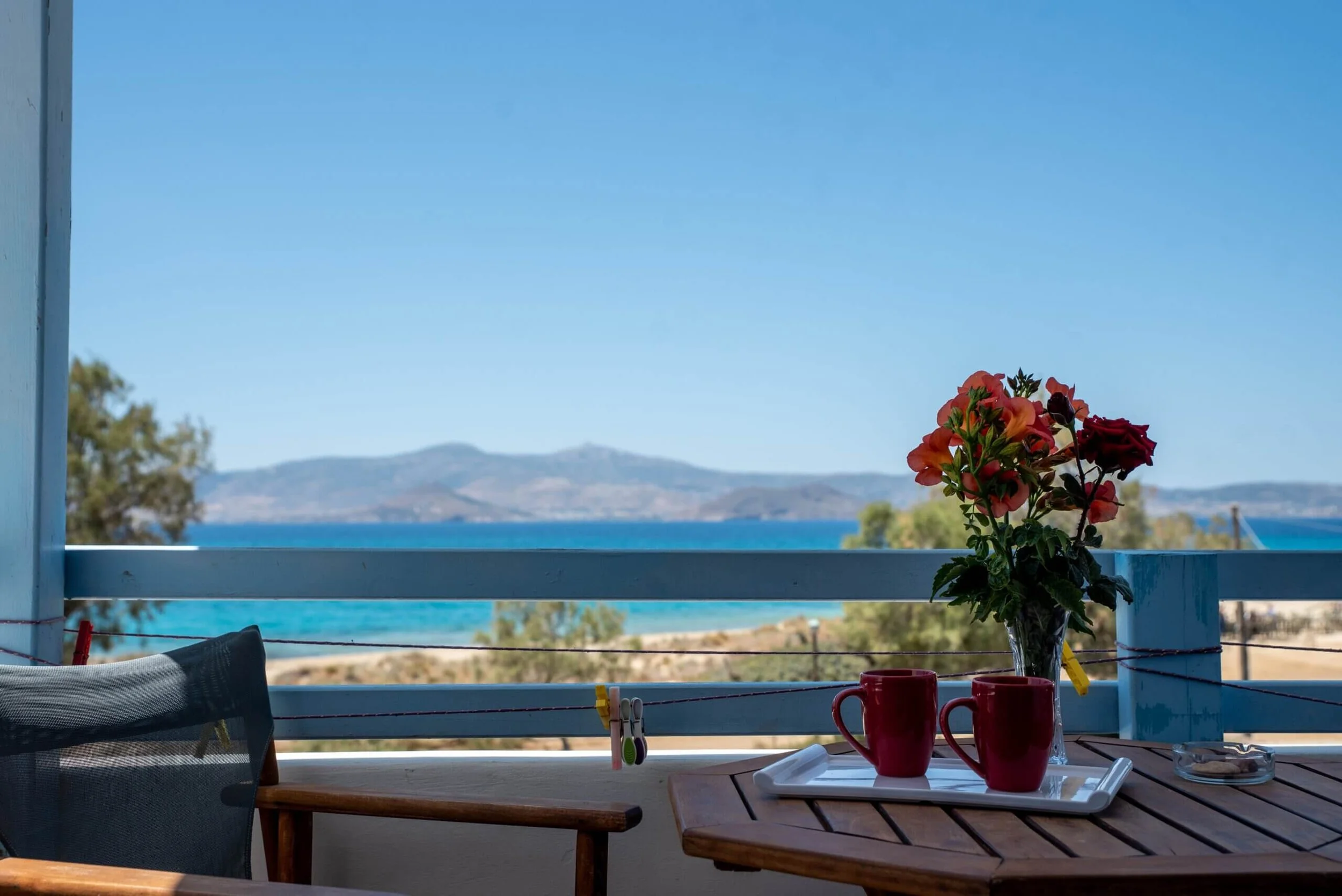 A balcony with a table holding a tray with two red mugs and a vase of colorful flowers. The view shows water and distant mountains under a clear blue sky.