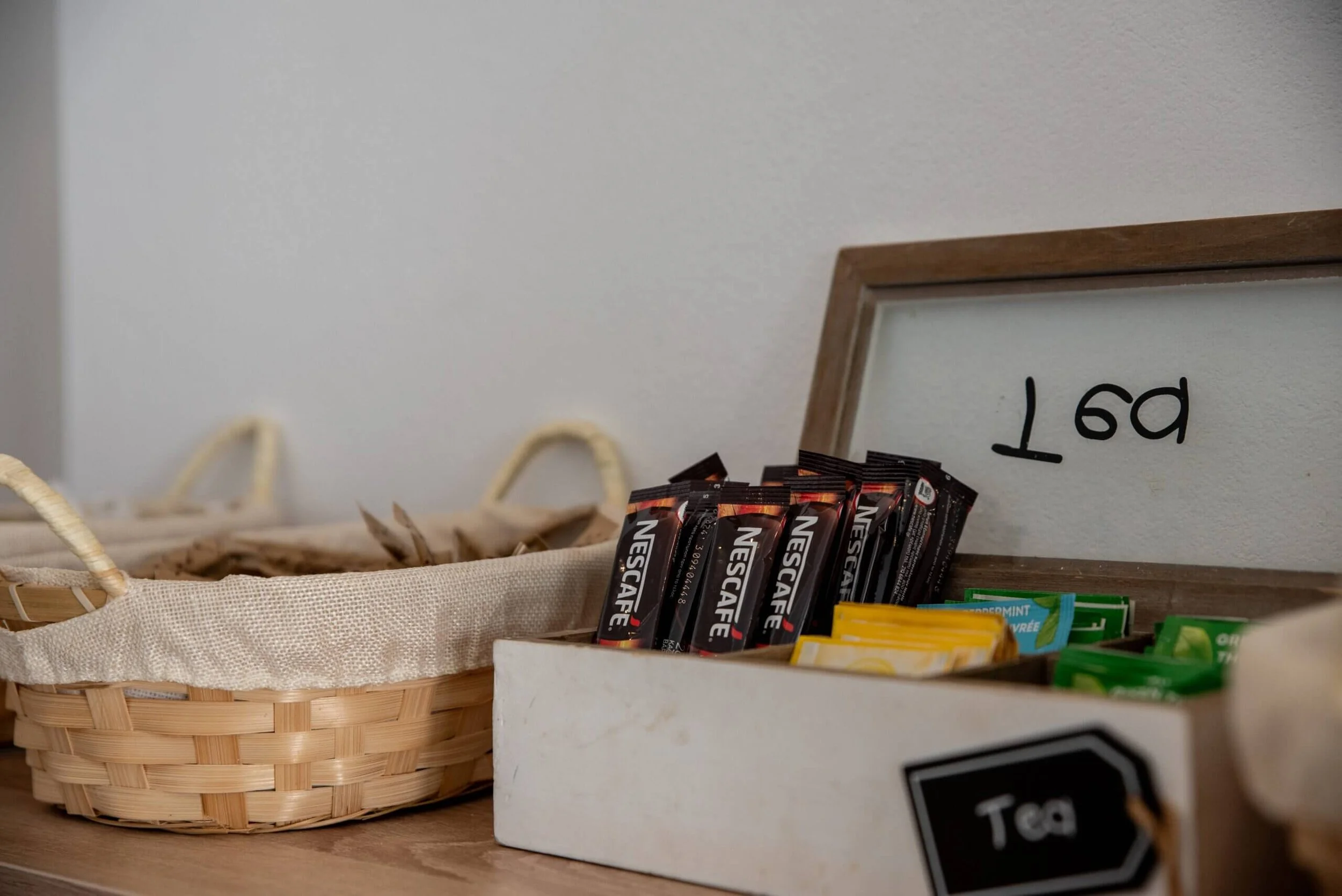 A basket with a cloth lining next to a white container filled with tea bags, including Nescafe coffee sticks and some tea packets, with a framed sign reading 'Tea' in the background.
