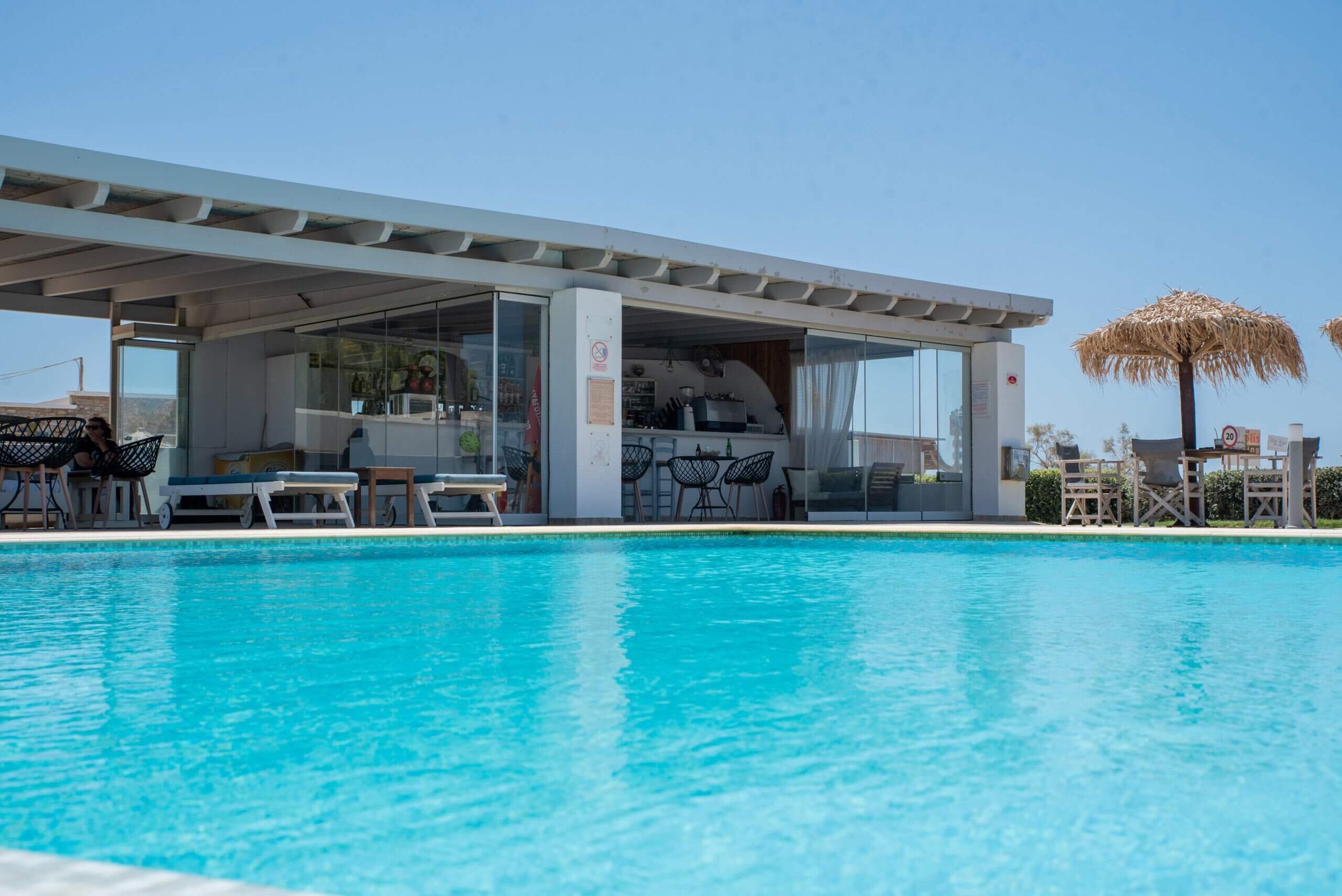 Poolside bar and seating area with tables and chairs, some with umbrellas, overlooking a swimming pool under a clear blue sky.