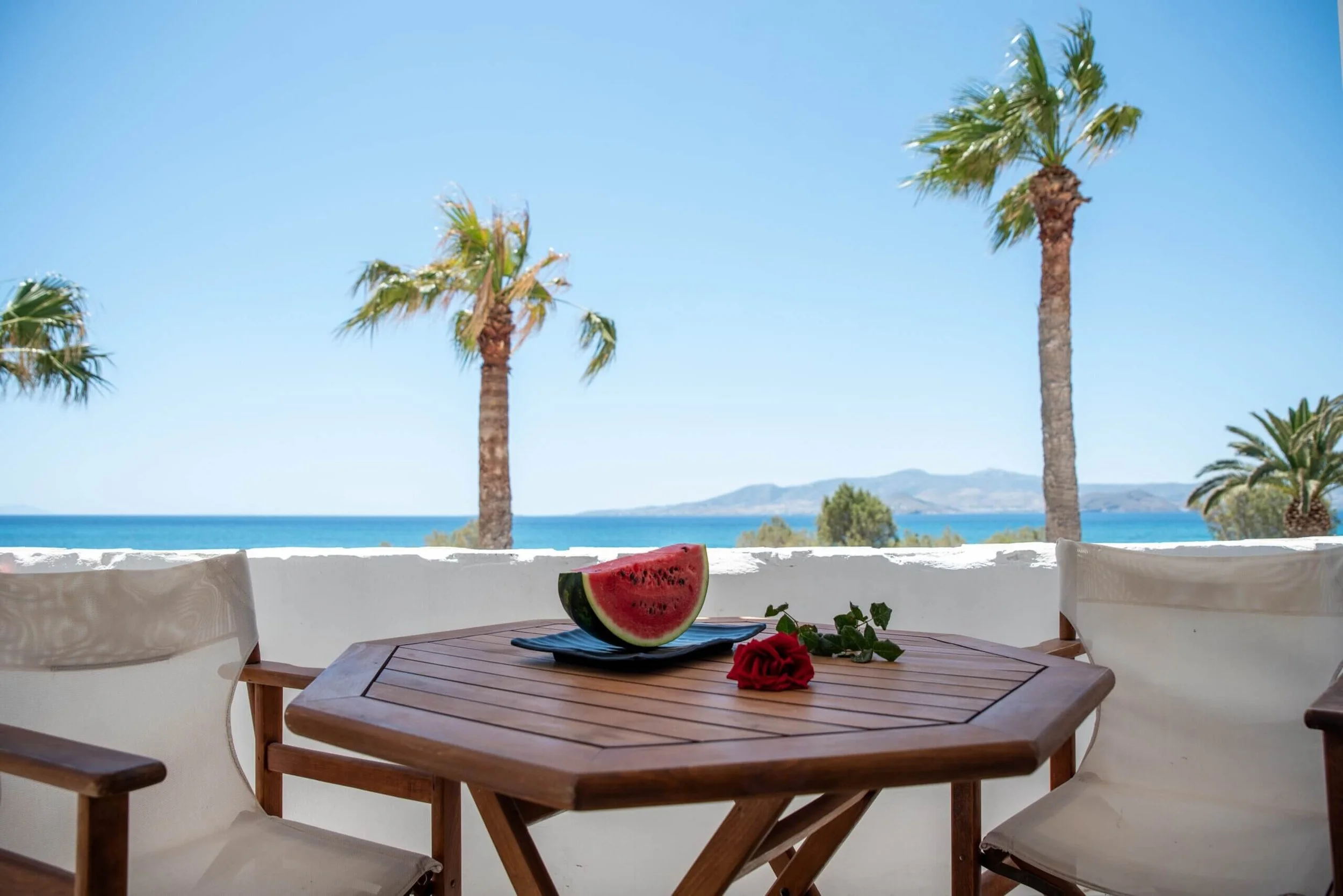 A seaside patio with a wooden table and two chairs. On the table, there is a slice of watermelon and a red rose. The background features a white wall, palm trees, a blue ocean, and distant hills under a clear sky.