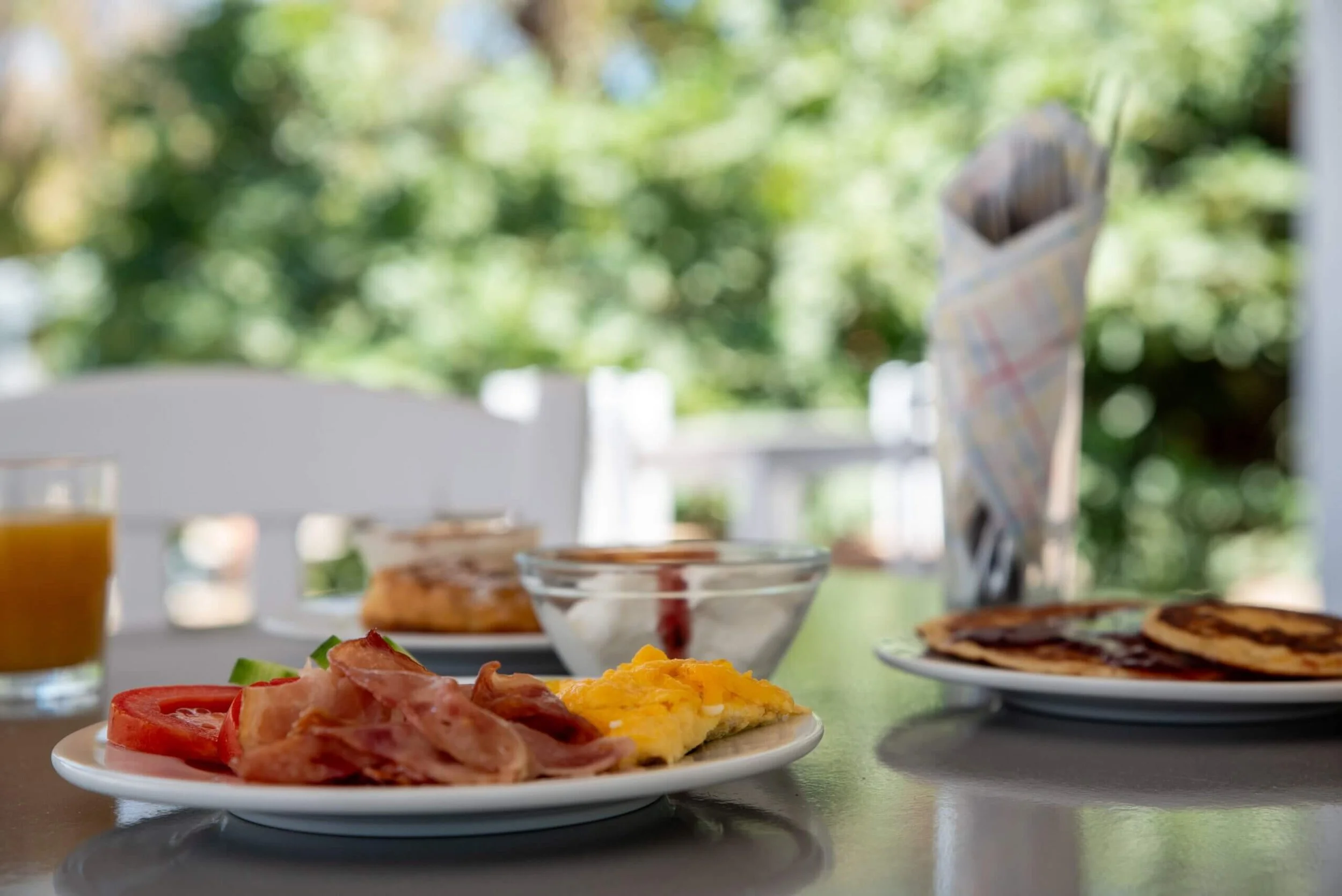 Breakfast table with bacon, scrambled eggs, tomato, pancakes, and juice seen in front of a blurred outdoor background.