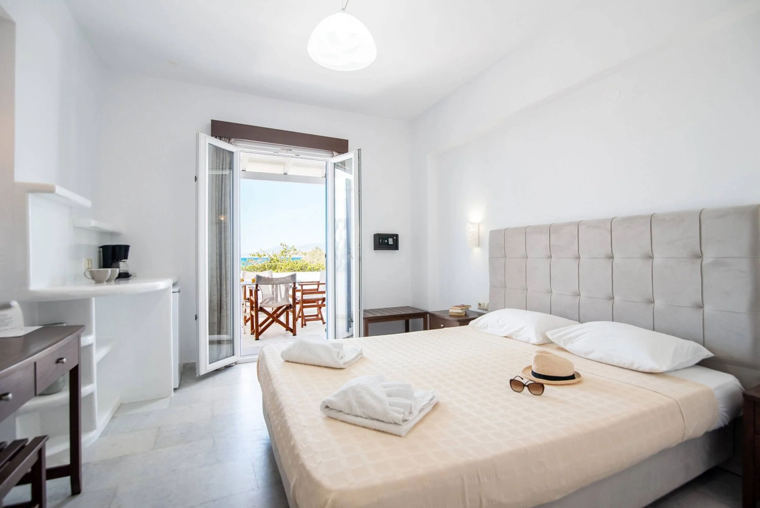 A bright hotel room with a balcony view, featuring a large bed with a beige headboard, towels, a hat, and sunglasses on it, a nightstand with books, and an open door leading to a small balcony with outdoor furniture and a view of greenery and the oce