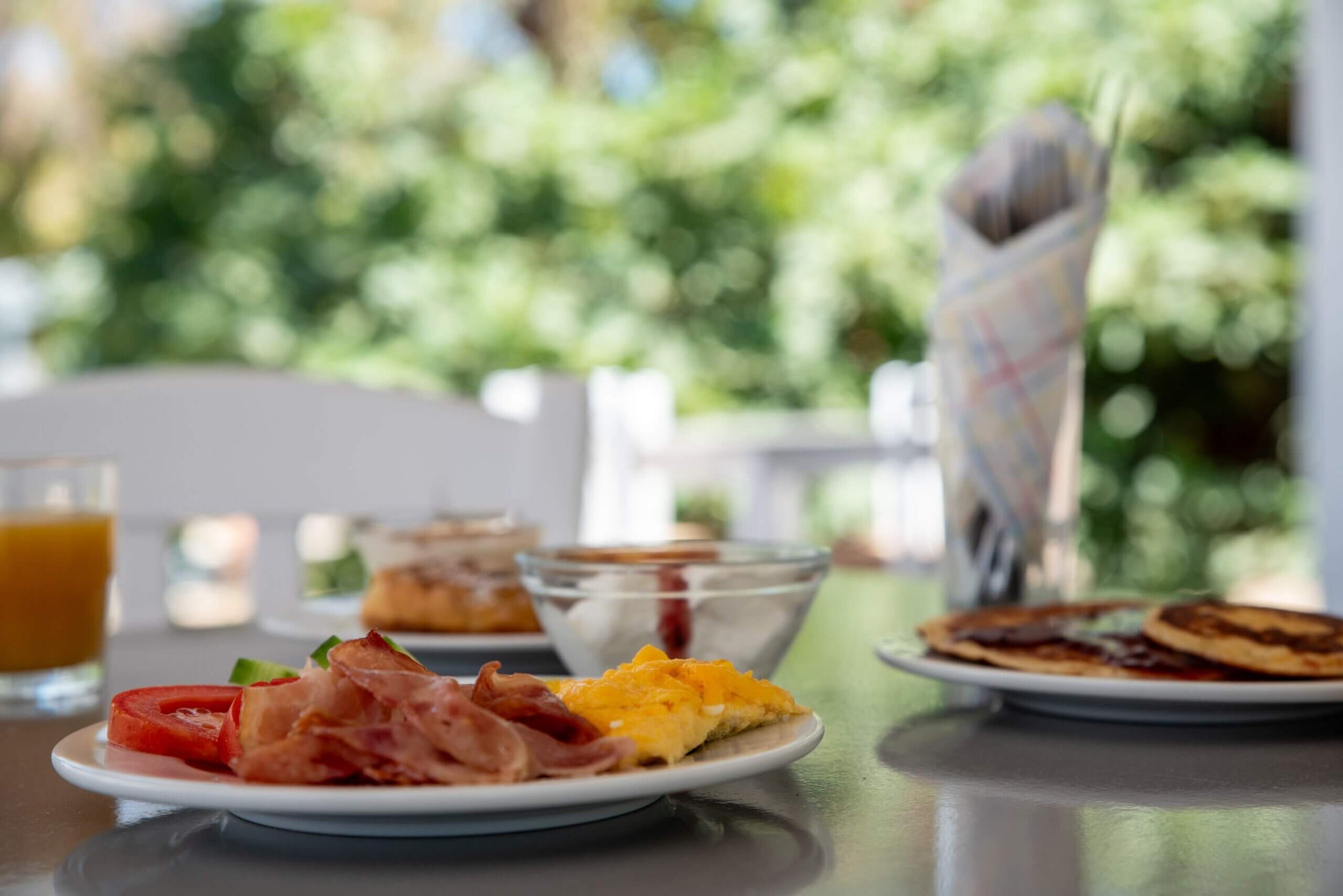 Breakfast on a table with plates of bacon, scrambled eggs, pancakes, a bowl of yogurt, a glass of orange juice, and a glass of tea against a background of green trees.