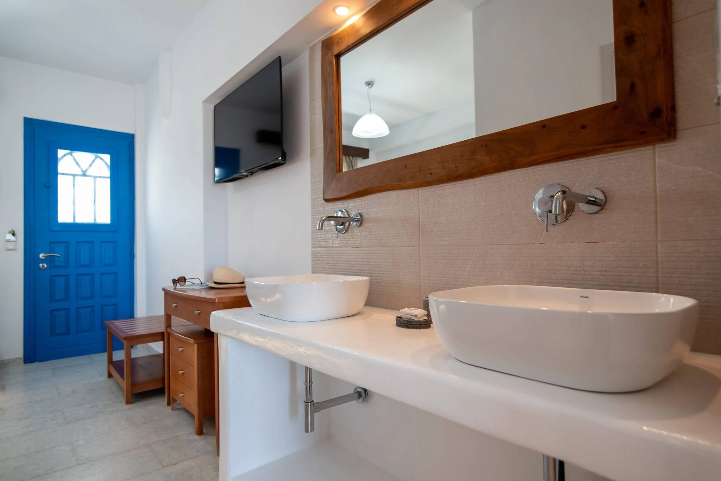 Double sink bathroom with white vessel sinks, a wooden-framed mirror, and a wall-mounted TV. Blue door and a small wooden table with a pair of sunglasses and a hat.