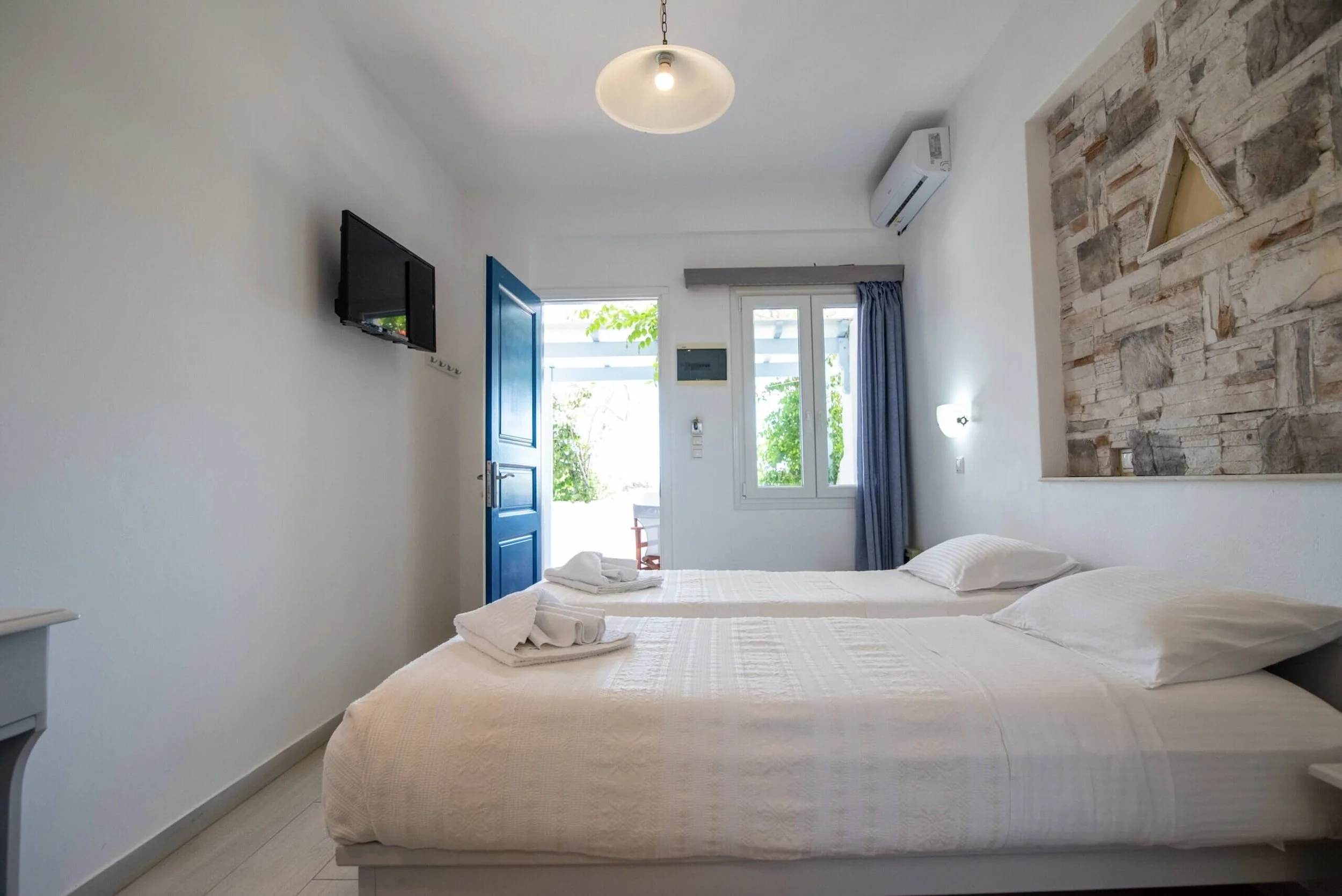 A bedroom with two white beds, a blue door open to outside, two windows with blue curtains, a wall-mounted TV, a stone accent wall, and an air conditioner.