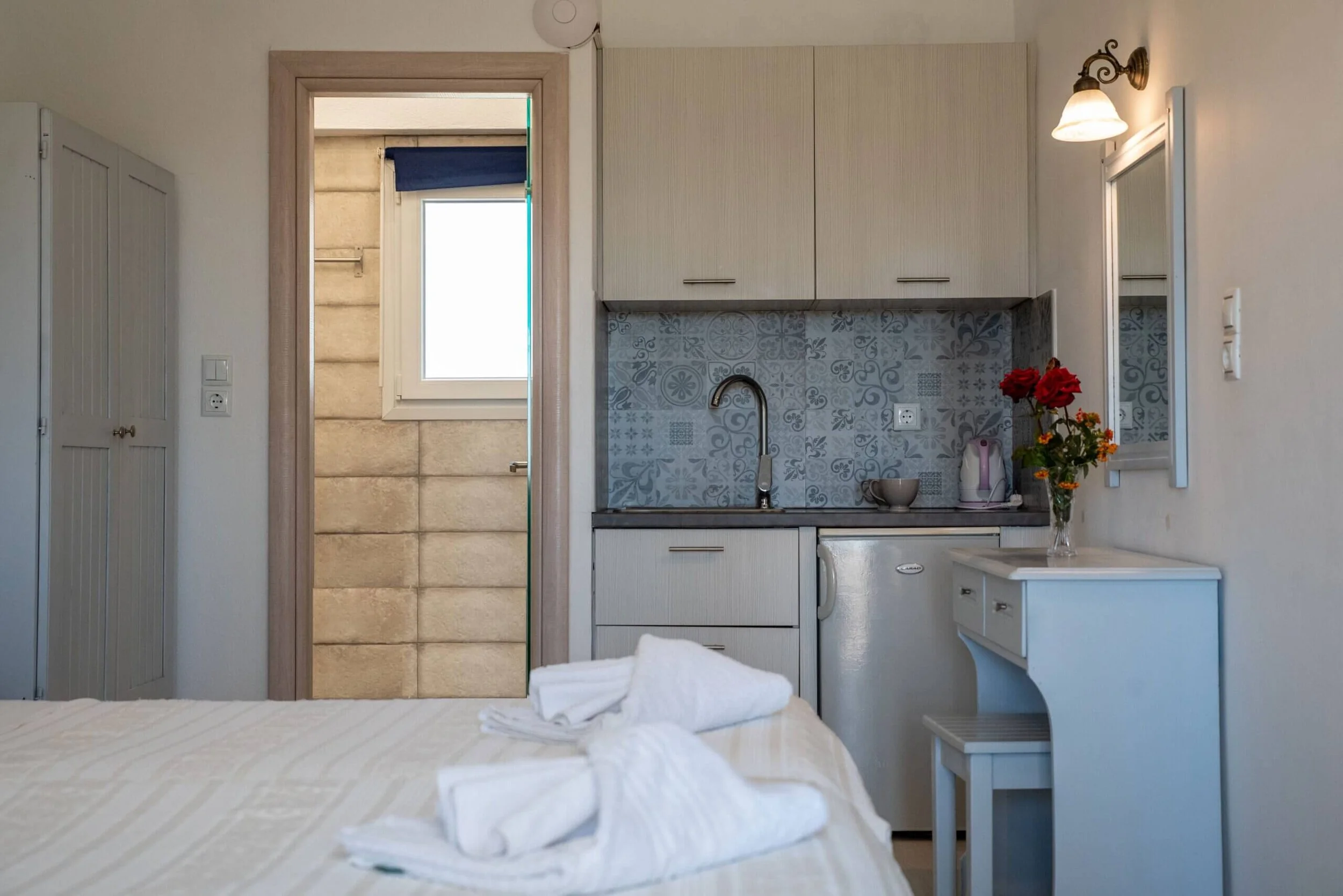 A small kitchenette in a compact living space with a sink, microwave, and refrigerator, decorated with a vase of red flowers, with a door leading to a bathroom shower area with a window.