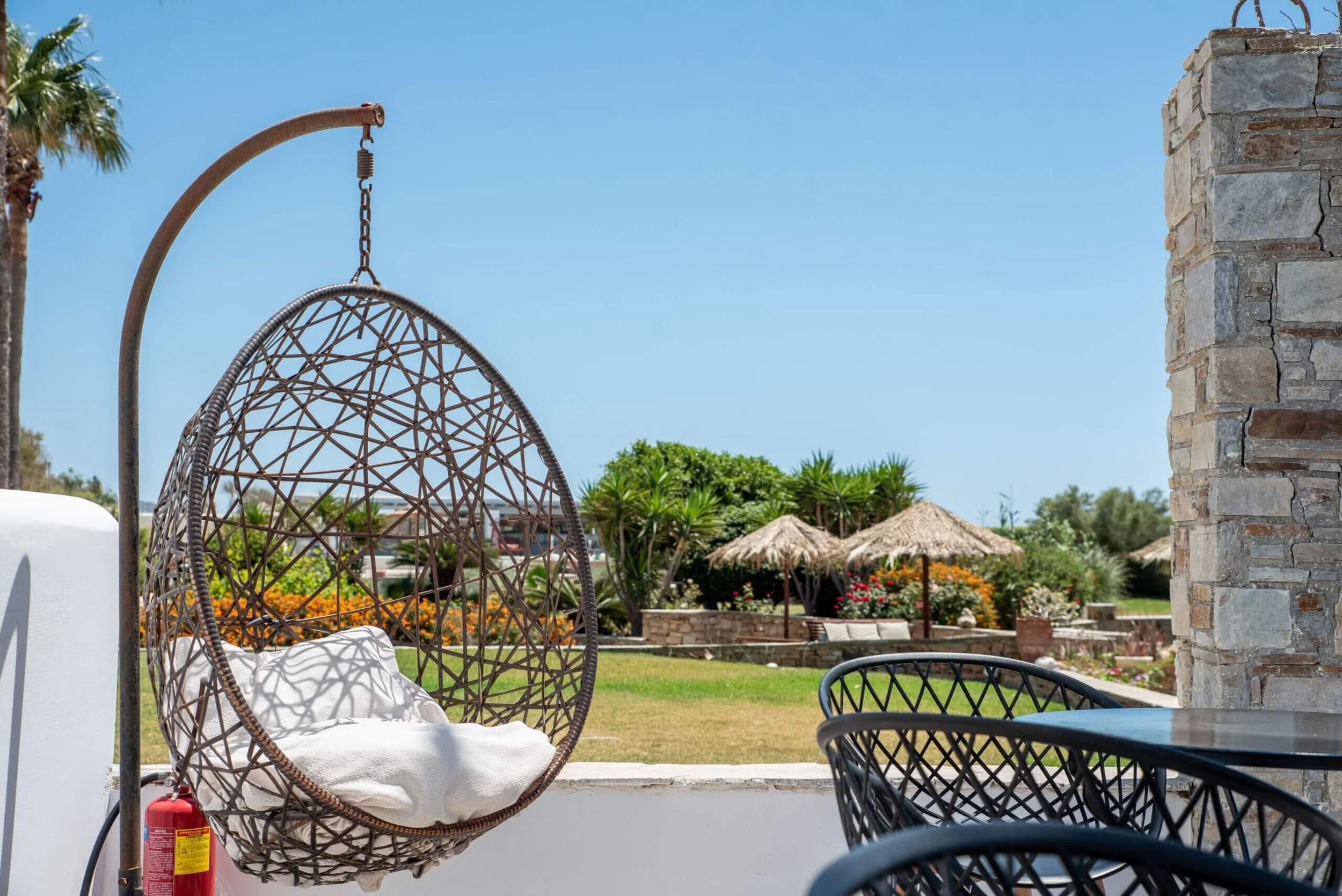Outdoor patio area with hanging wicker egg chair with white cushion, black metal chairs and table, stone pillar, lush green landscaping, thatched umbrellas, and clear blue sky.