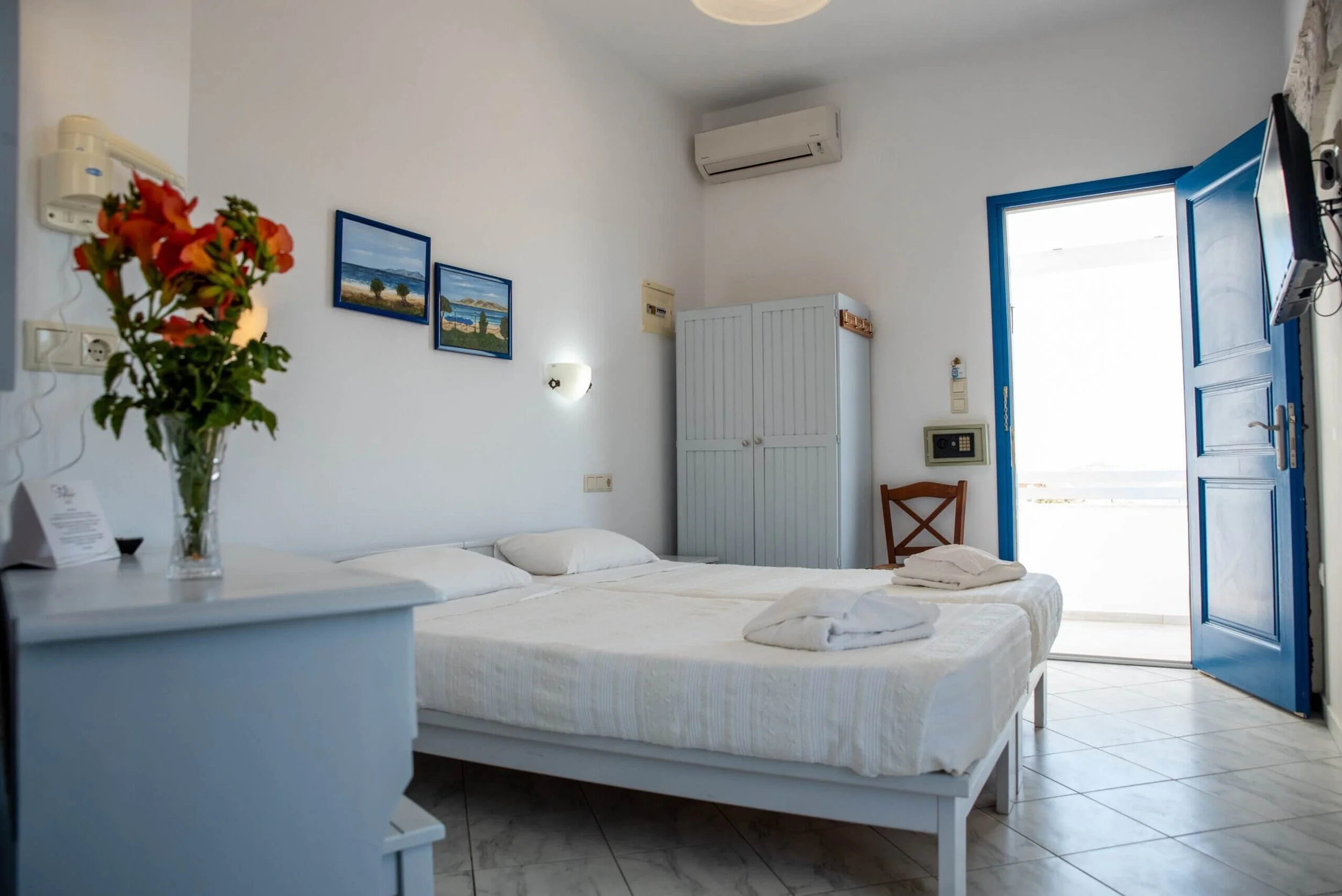 Hotel room with white walls, a bed with white sheets, a bedside table with a vase of flowers, paintings on the wall, a wooden chair, a white wardrobe, a safe on the wall, an air conditioner, and a door leading outside.