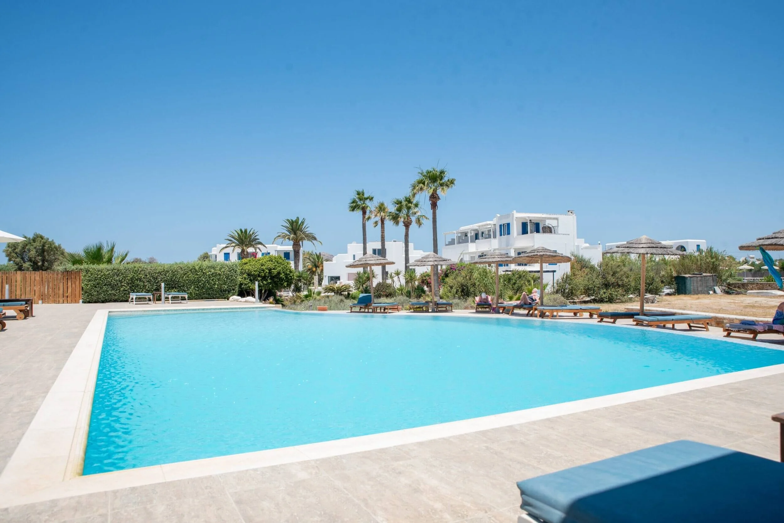 A swimming pool in a sunny resort with sun loungers and umbrellas, surrounded by white buildings, palm trees, and a clear blue sky.