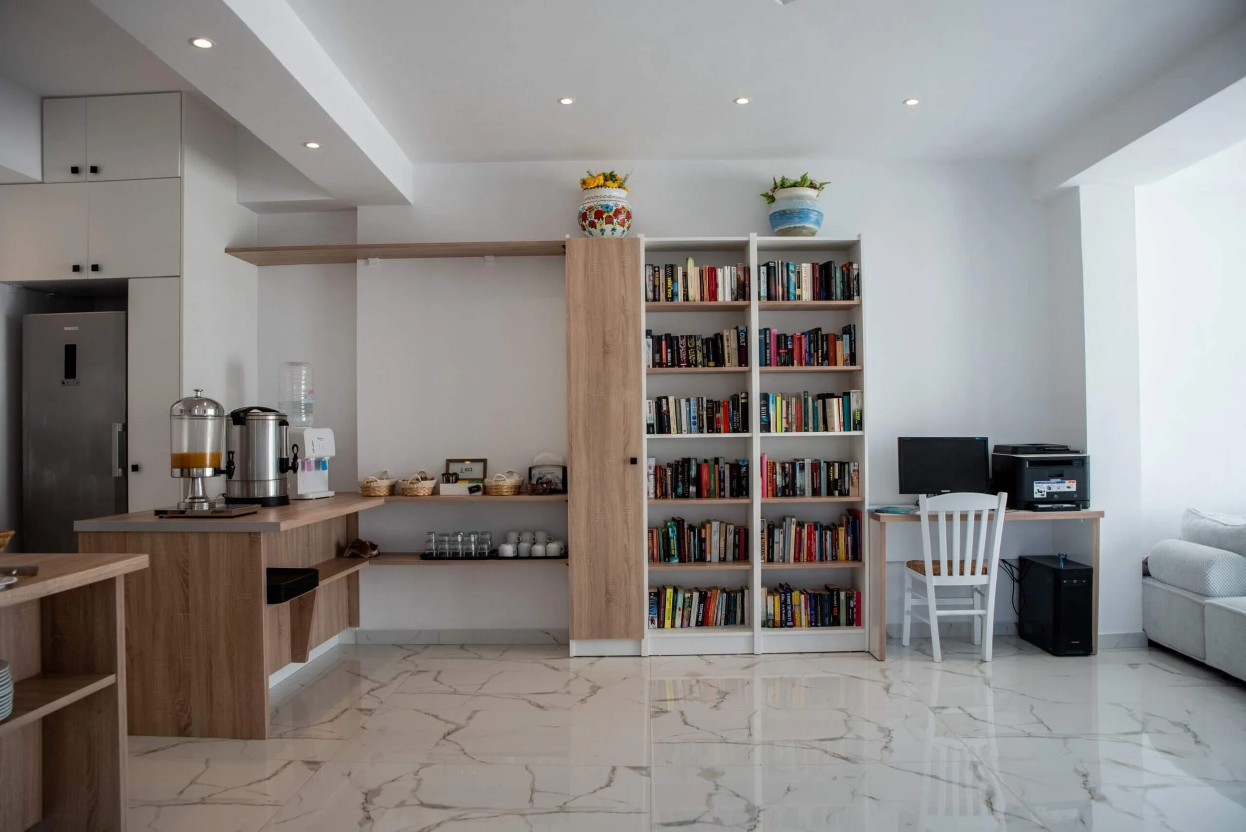 Living room with a white bookshelf filled with books, a desk with a computer and printer, and a white sofa, featuring white walls and marble flooring.