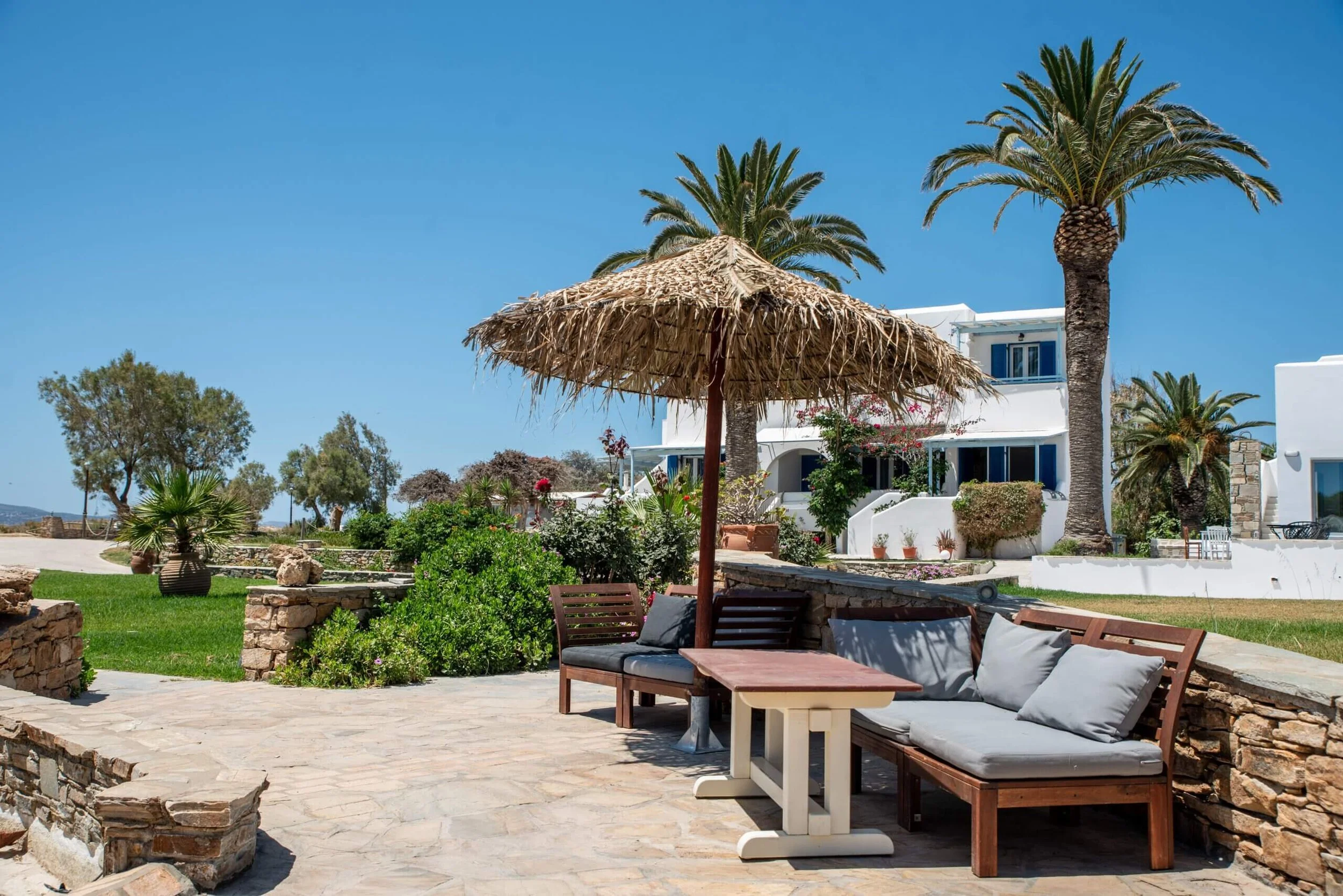 Outdoor patio with wooden benches, gray cushions, a small wooden table, and a thatched umbrella. There are palm trees, colorful bushes, and white buildings in the background under a clear blue sky.