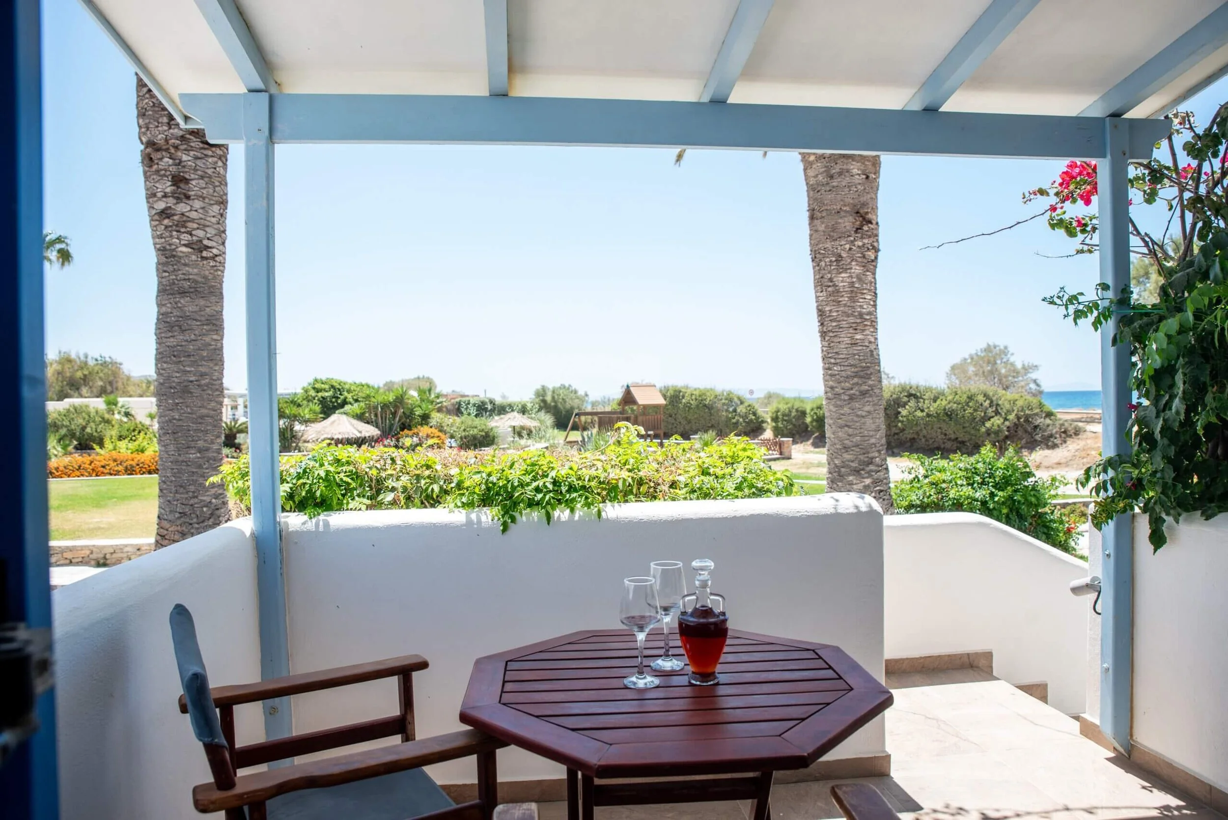 A patio with a small wooden table set with a wine bottle and two wine glasses, surrounded by lush greenery and palm trees, overlooking a bright, sunny landscape.
