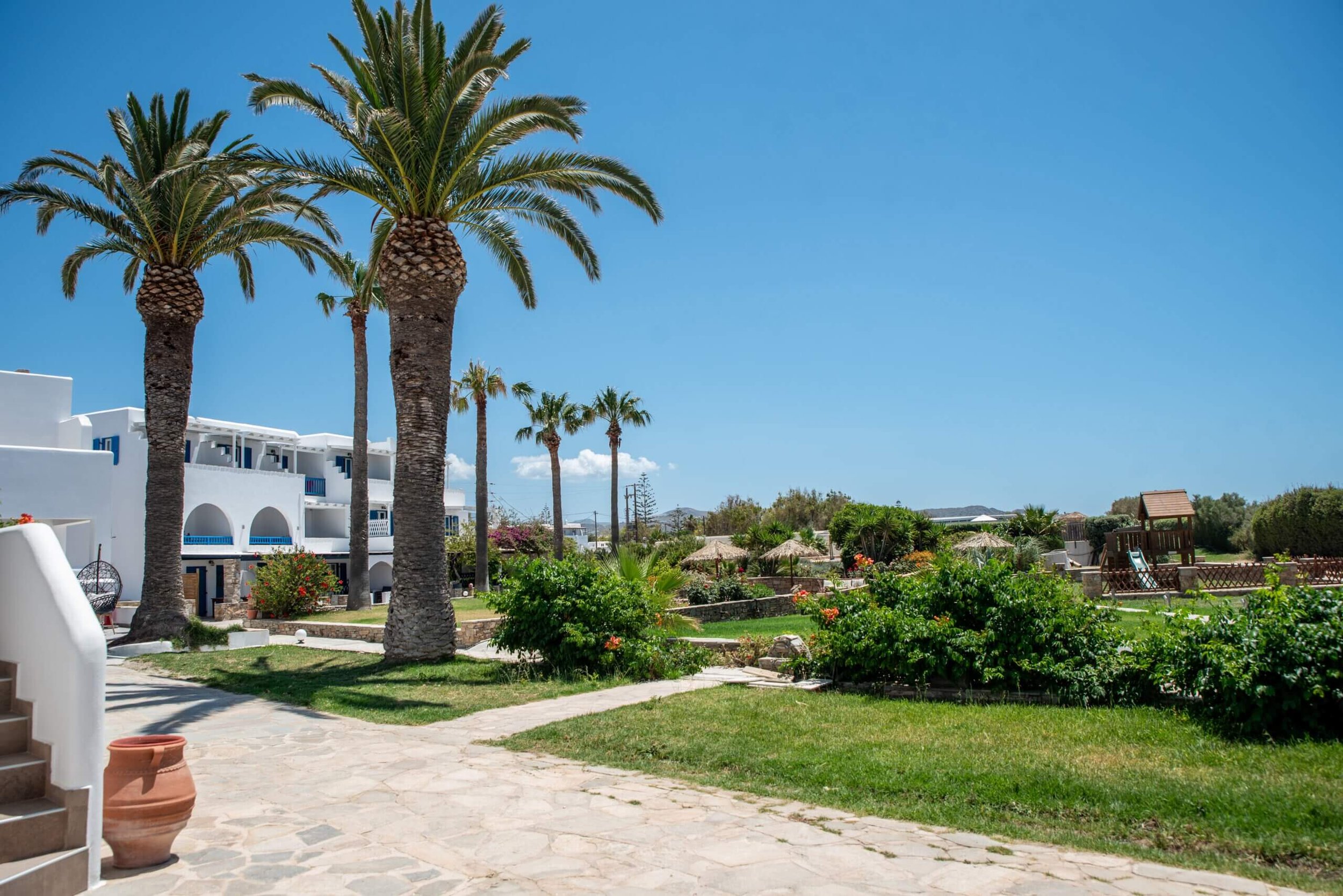 Bright sunny day at a tropical resort with white buildings, tall palm trees, green bushes, and a children's playground with a slide and houses in the background.