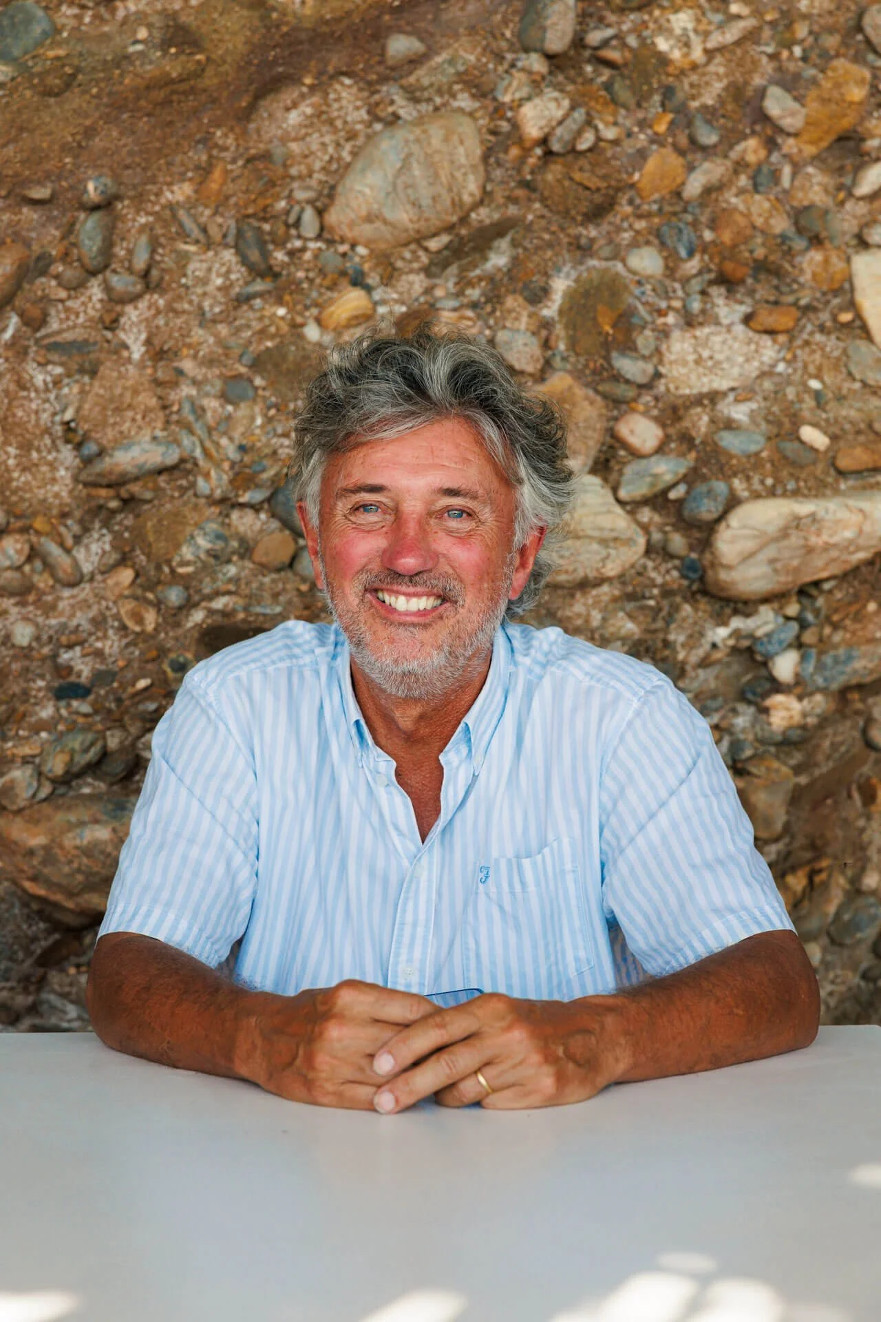 A smiling middle-aged man with gray hair and a beard, wearing a blue and white striped shirt, sitting at a table in front of a rocky background.