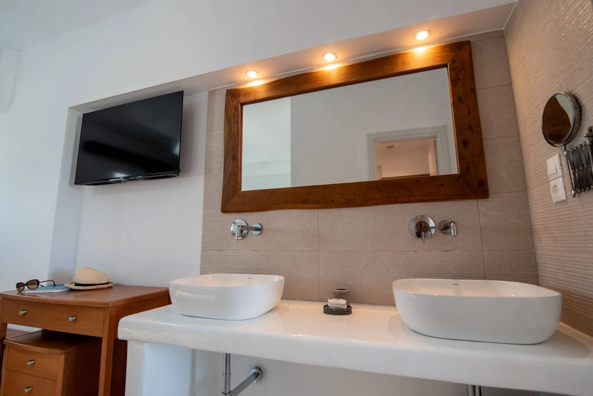 Bathroom with two vessel sinks on a white countertop, a large wooden-framed mirror, wall-mounted faucets, a flat-screen TV on the wall, a wooden cabinet with hat and sunglasses, a small mirror and toiletries on the side, and beige tiled walls.