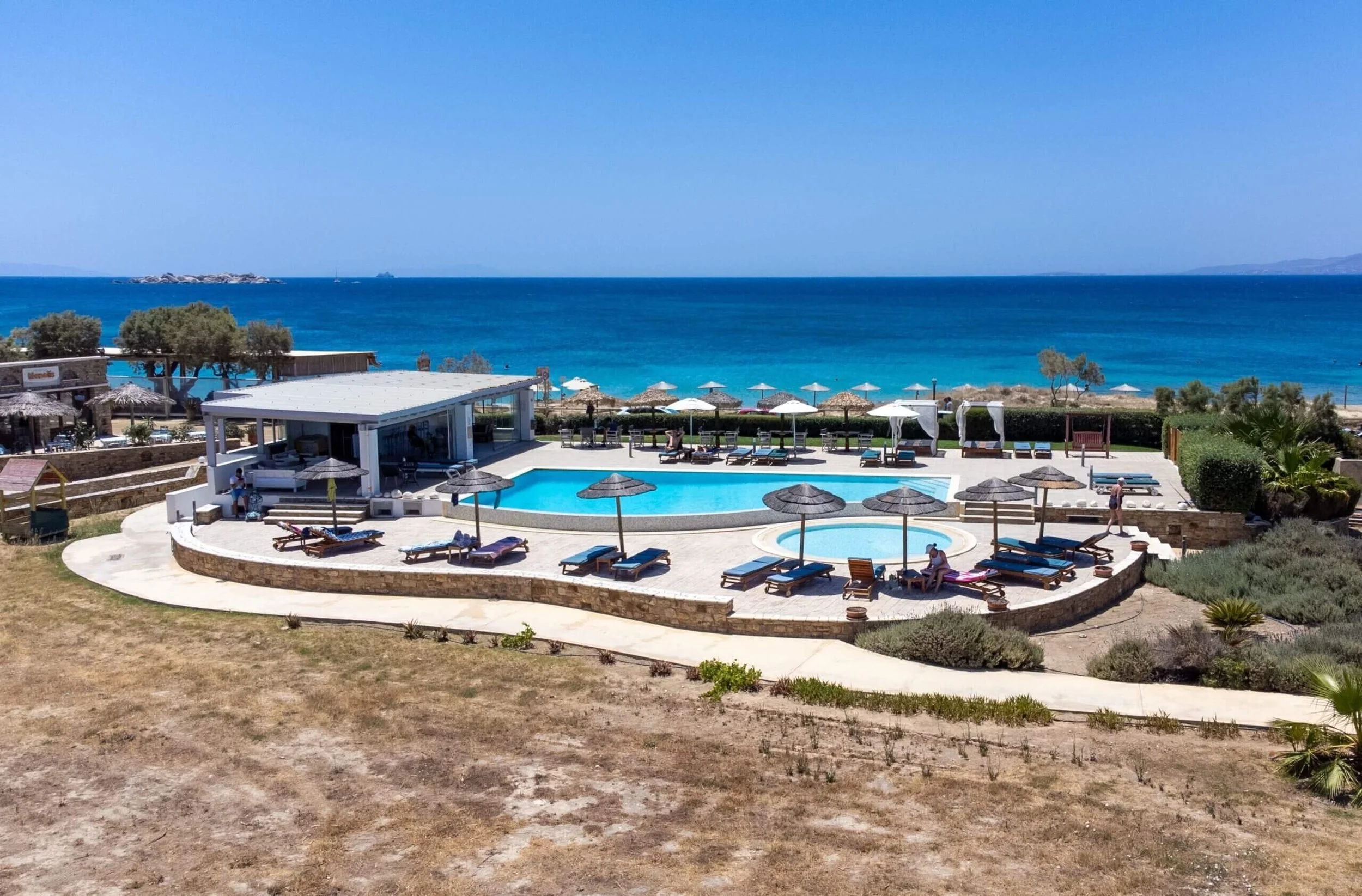 A seaside resort with a swimming pool and hot tub, surrounded by lounge chairs and umbrellas, overlooking the ocean with a clear blue sky.