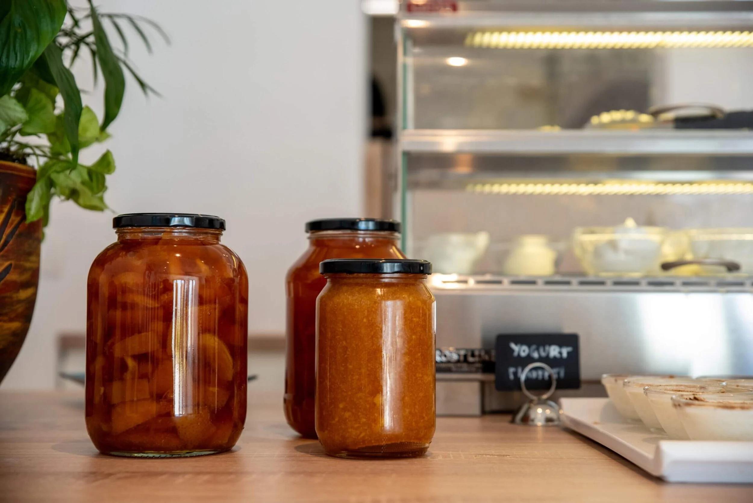 Three jars of homemade preserves or jams on a wooden surface next to a plant and a refrigerated display case with bowls of yogurt in the background.
