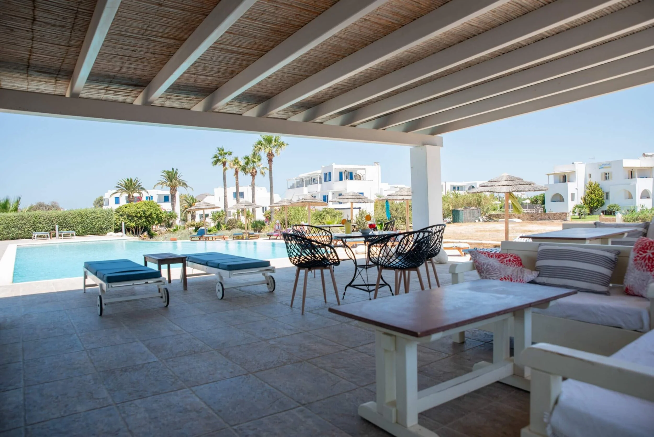 Outdoor patio with pool, lounge chairs, and seating under a wooden pergola, overlooking white buildings and palm trees on a clear sunny day.