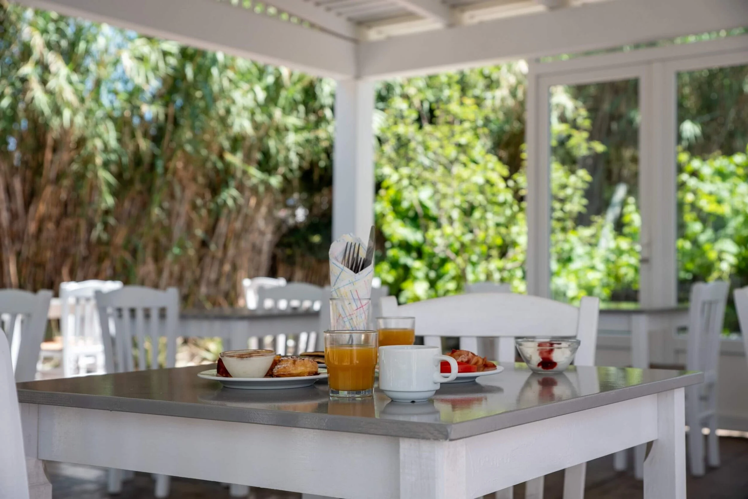 Breakfast table set outdoors with orange juice, coffee, cereal, toast, and fruit, surrounded by white chairs and greenery.