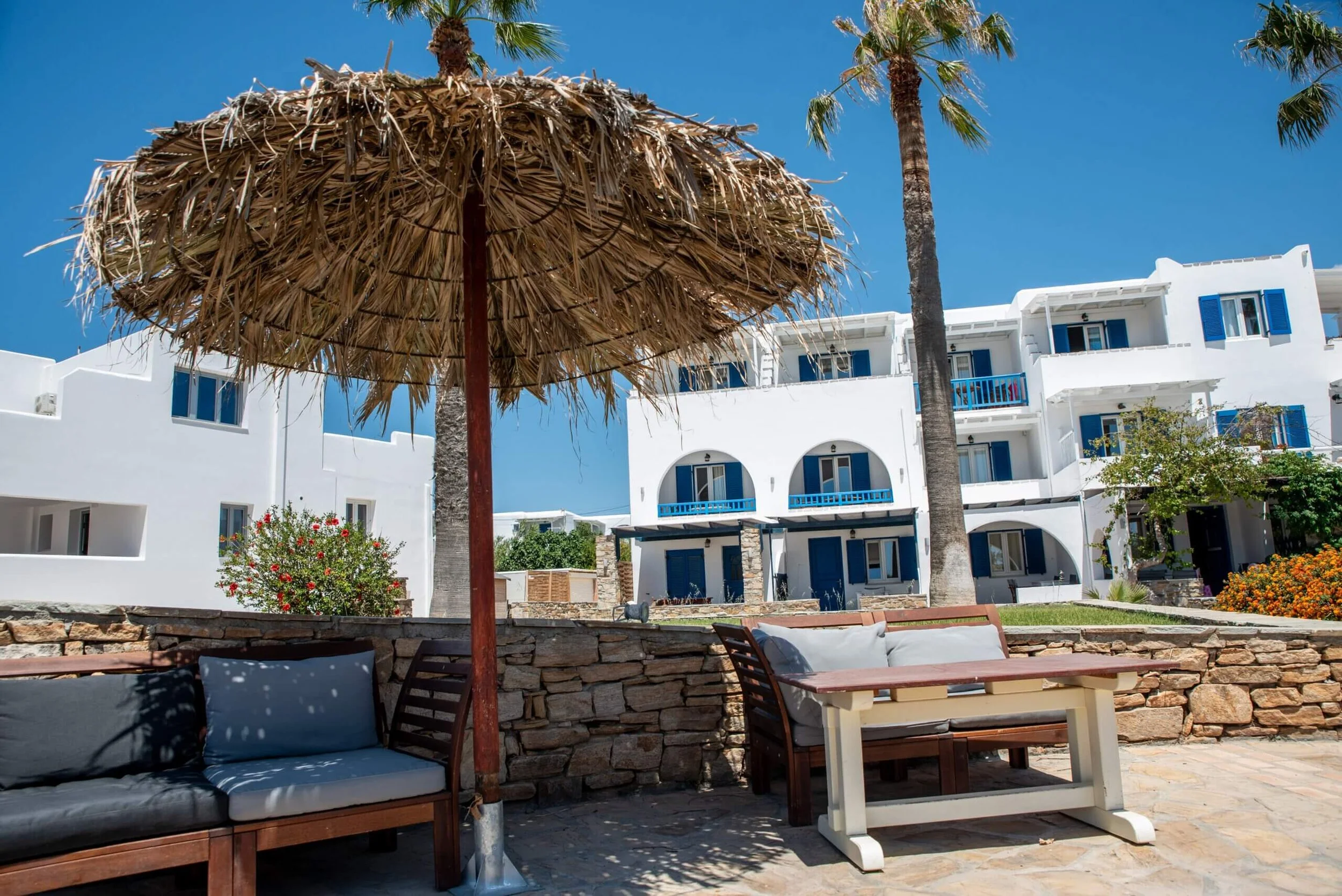 Outdoor seating area with benches and a straw umbrella in front of white buildings with blue accents, palm trees, and flowering plants under a clear blue sky.