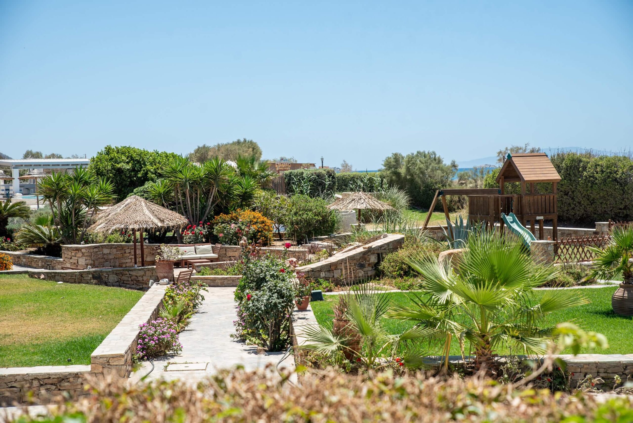 A lush outdoor garden with various plants, flowering bushes, and trees, featuring a stone pathway, a wooden playhouse with a slide, and a thatched umbrella providing shade over garden furniture on a sunny day.