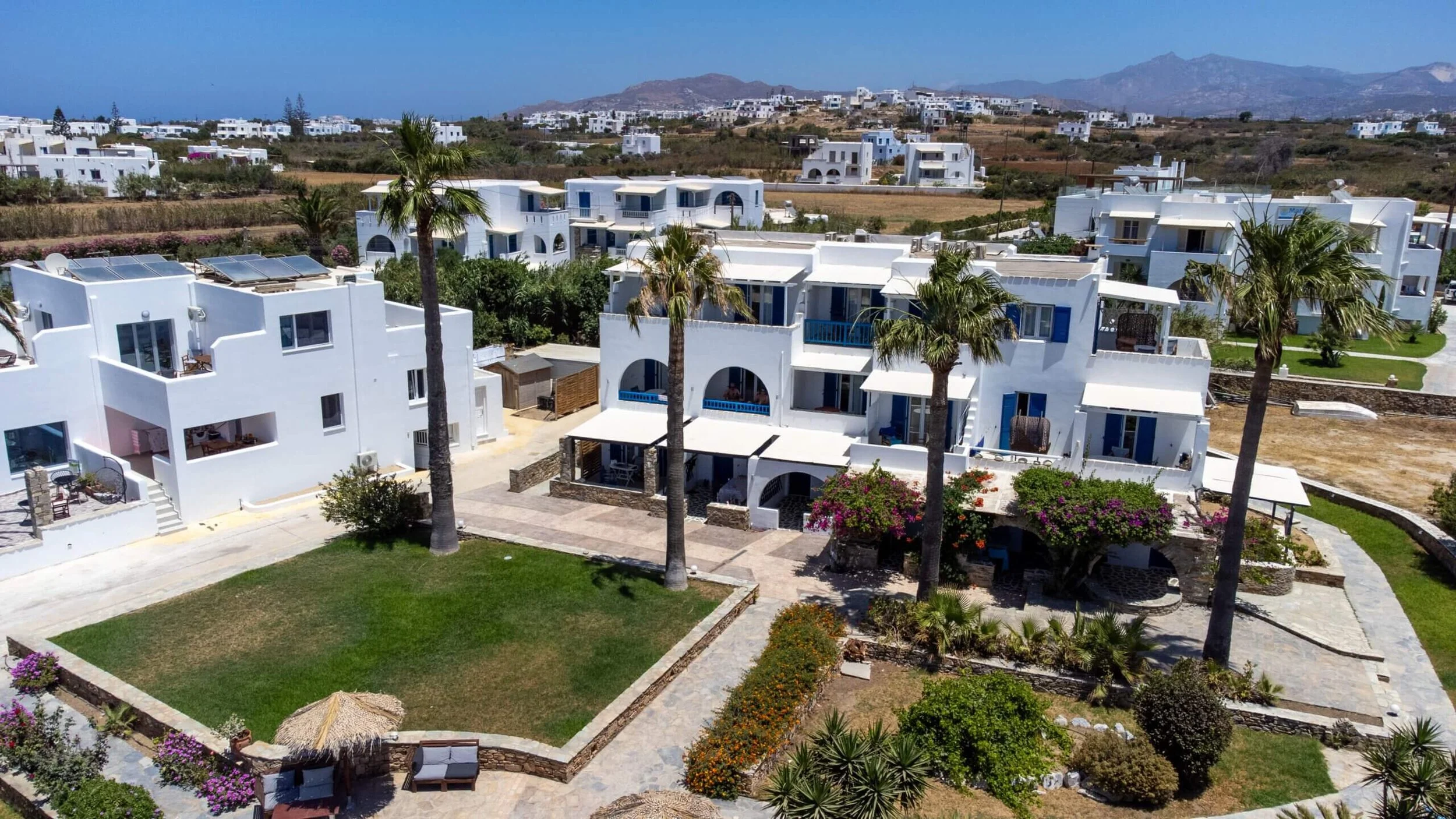 Aerial view of white Mediterranean-style residential buildings with blue accents, surrounded by palm trees and landscaped gardens, in a coastal area with mountains in the background.