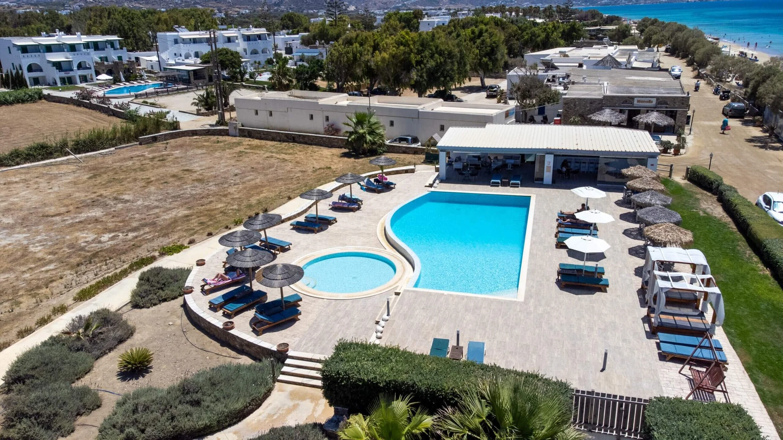 Aerial view of a hotel and pool area near a beach, featuring a large rectangular swimming pool, a smaller circular hot tub, sun loungers with umbrellas, a poolside bar or lounge, and a sandy beach with umbrellas and people near the ocean in the backg