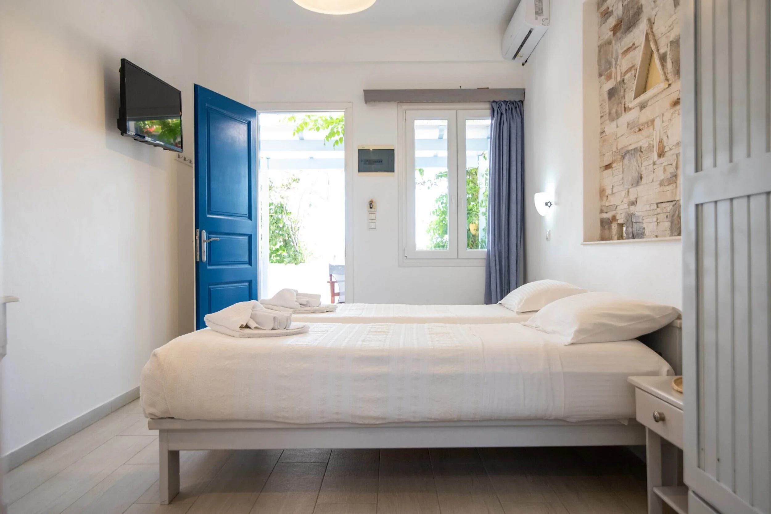 A bright hotel room with two double beds, white linens, and pillows. There are towels on the beds. The room has a blue door, a window with a view outside, and a wall-mounted TV. The decor includes white walls and a stone accent wall.