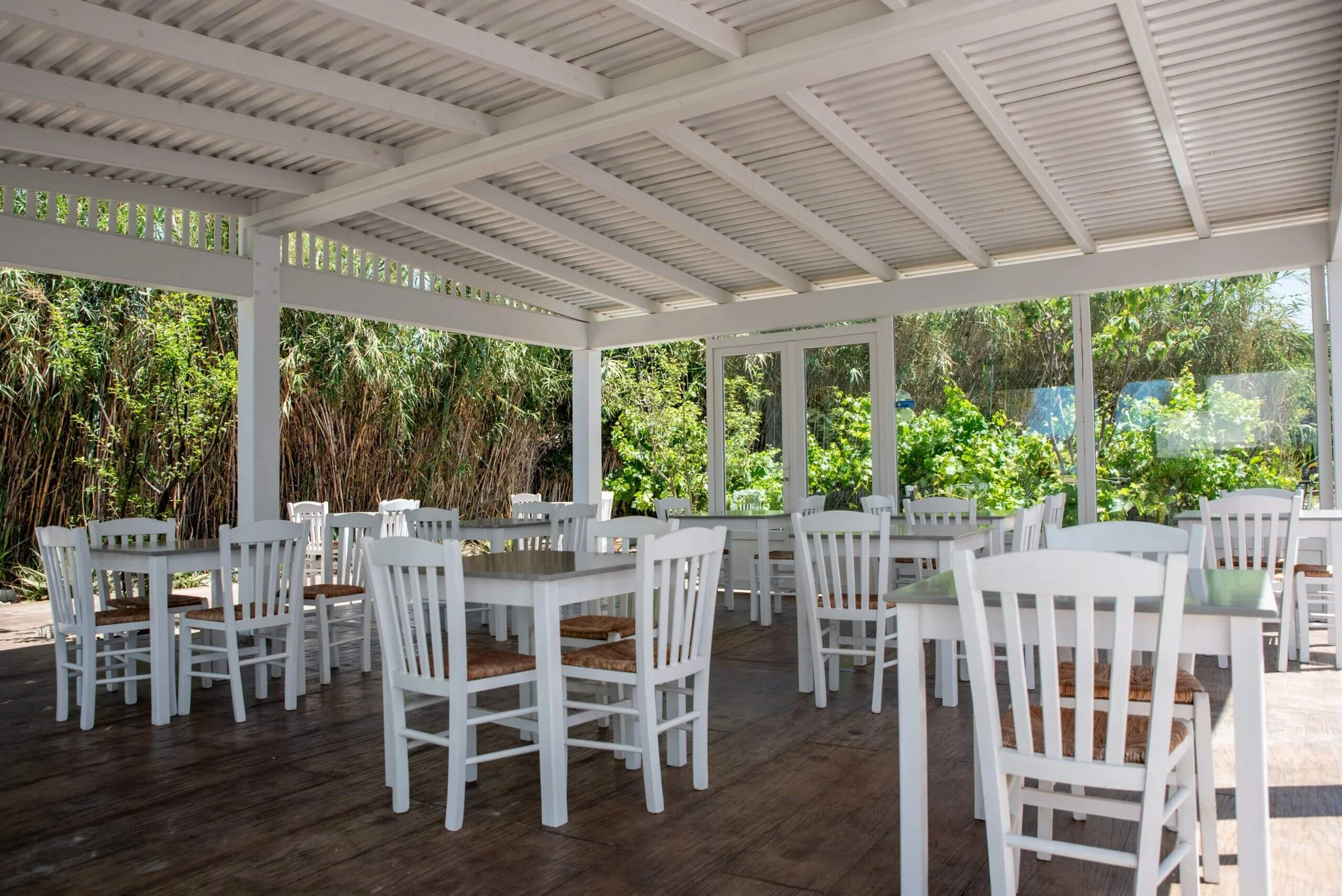 An outdoor patio area with white wooden chairs and tables under a covered roof, surrounded by greenery.