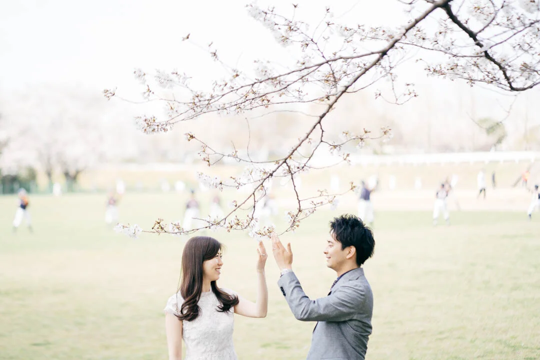 A man and woman stand under a cherry blossom tree, touching hands and smiling at each other, with a sports field and people in the background.