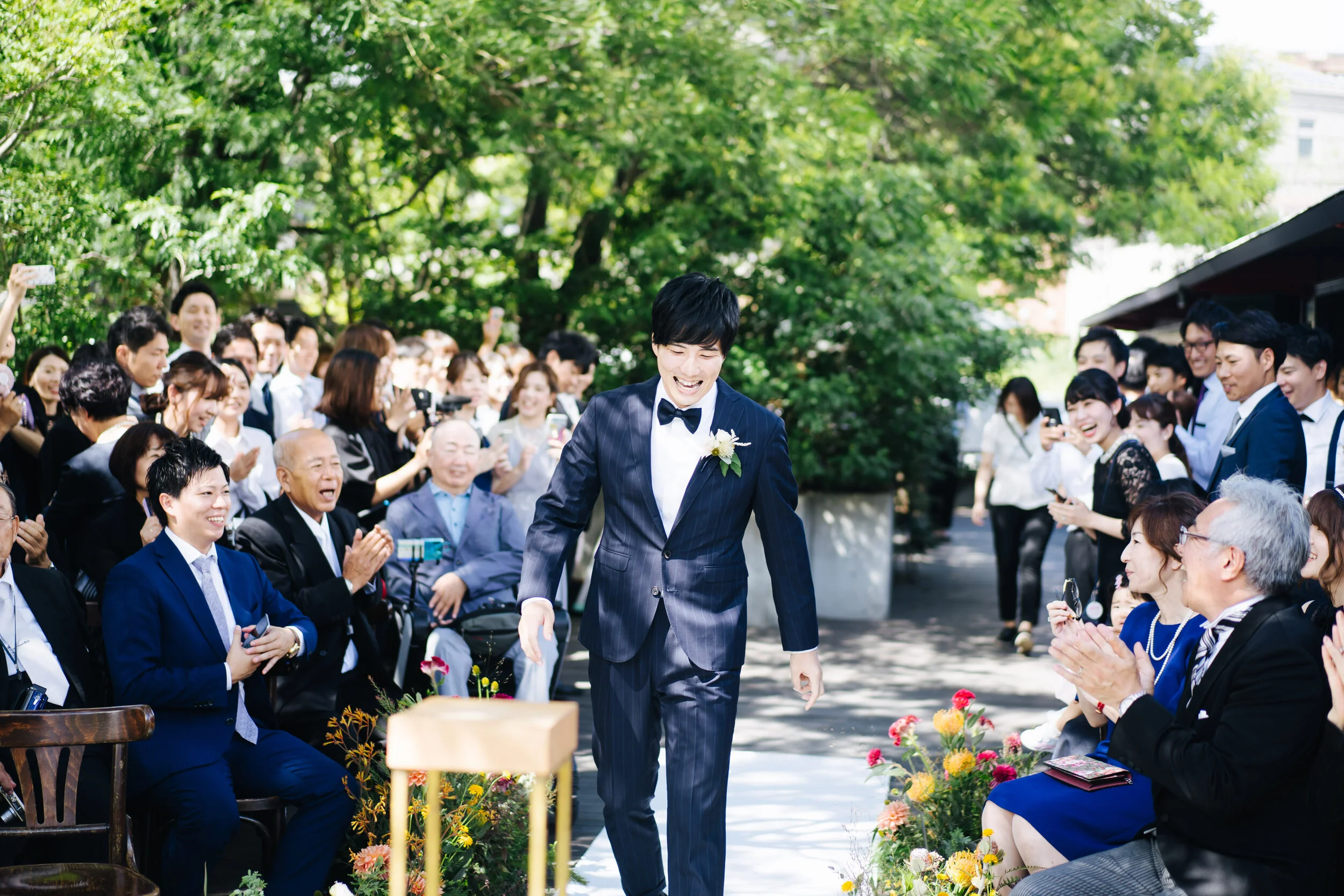 A groom in a dark striped suit and bow tie walking down the aisle at an outdoor wedding, surrounded by seated guests and green trees.