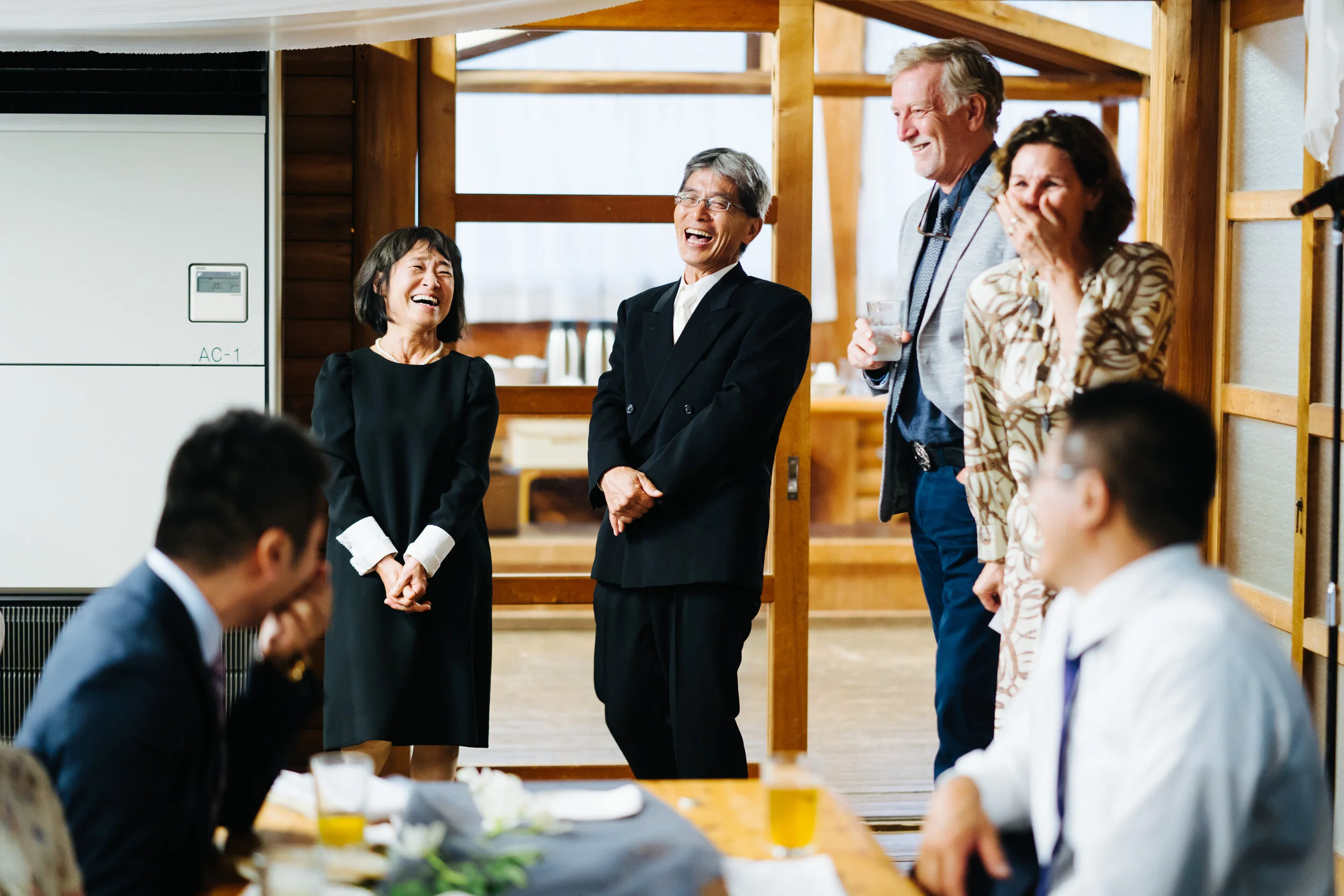 A group of adults at a social gathering, smiling and laughing while standing and sitting in a wooden indoor setting.