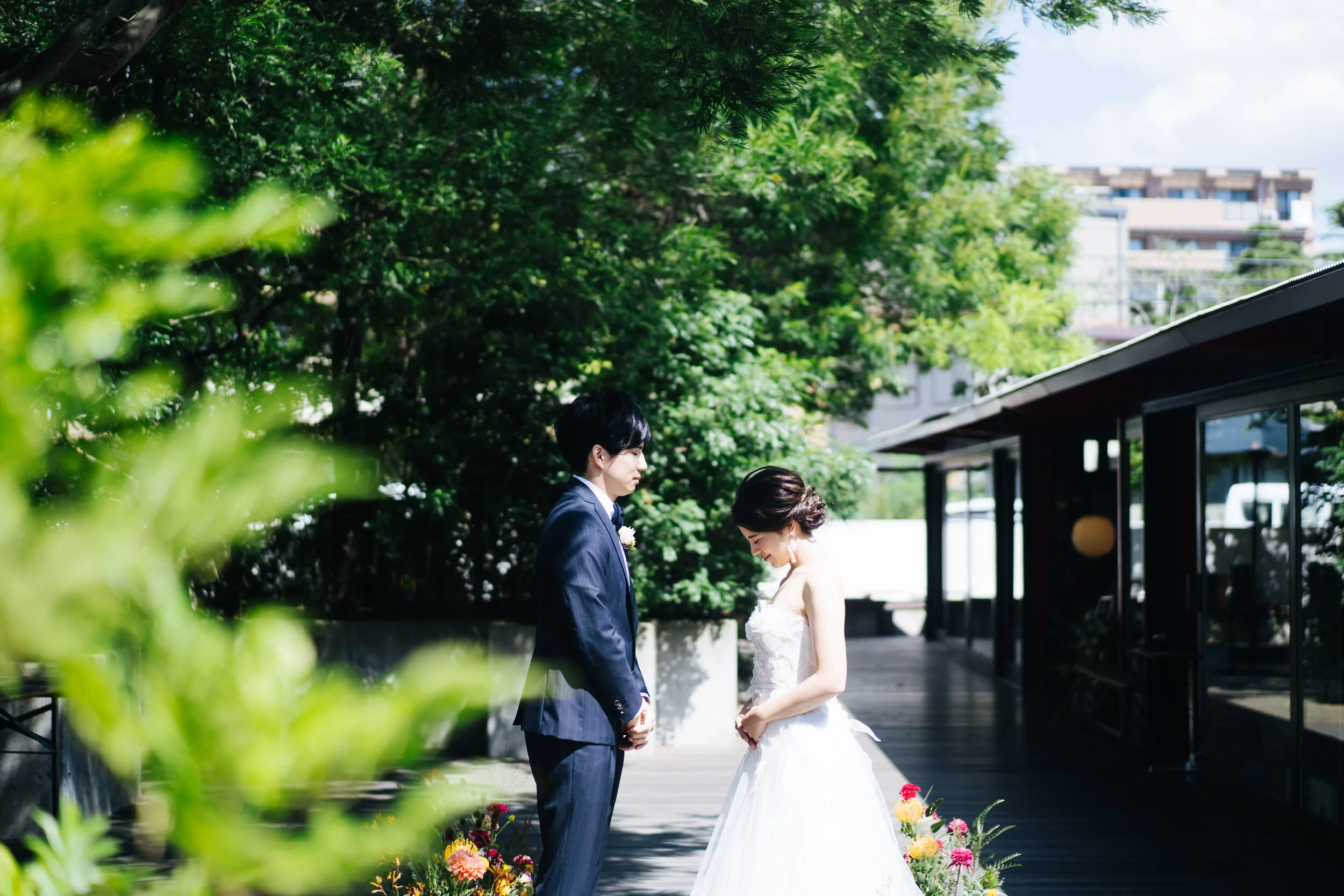 A bride and groom standing outdoors, holding hands, with greenery and trees around them on their wedding day.