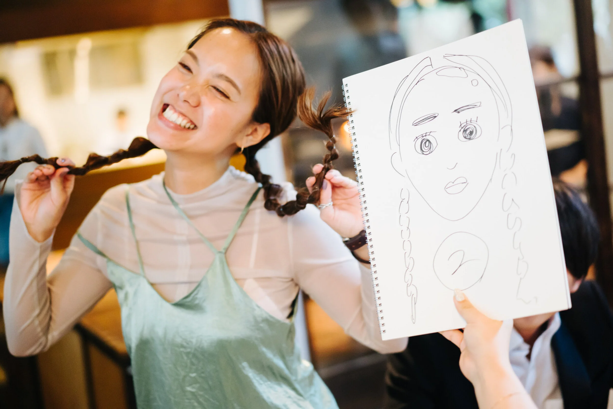 A smiling woman with braided hair holding a spiral notebook with a drawing of a person's face and upper body in front of her.