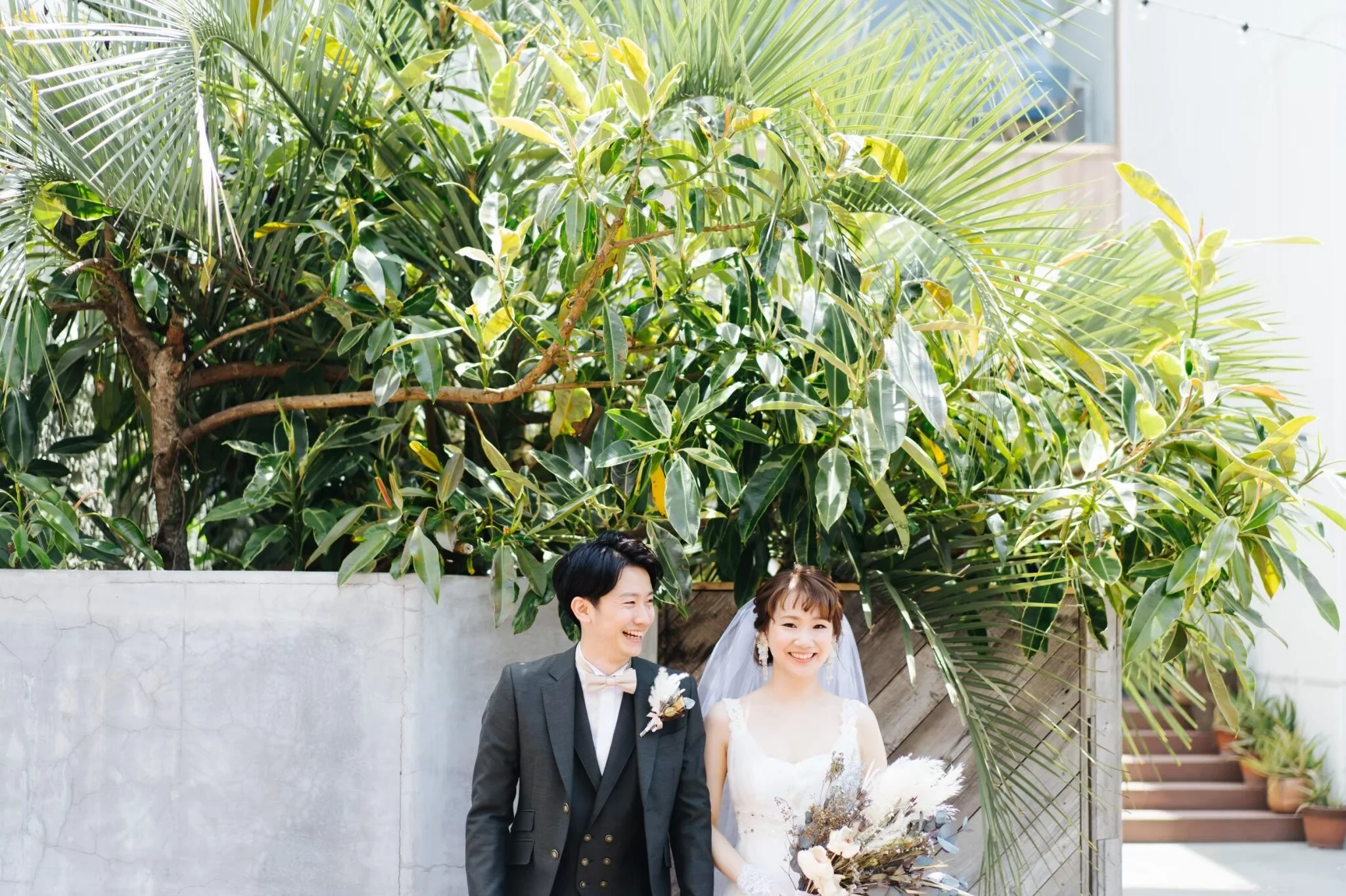 A newly married couple standing outside in front of lush green tropical plants, smiling happily on their wedding day.