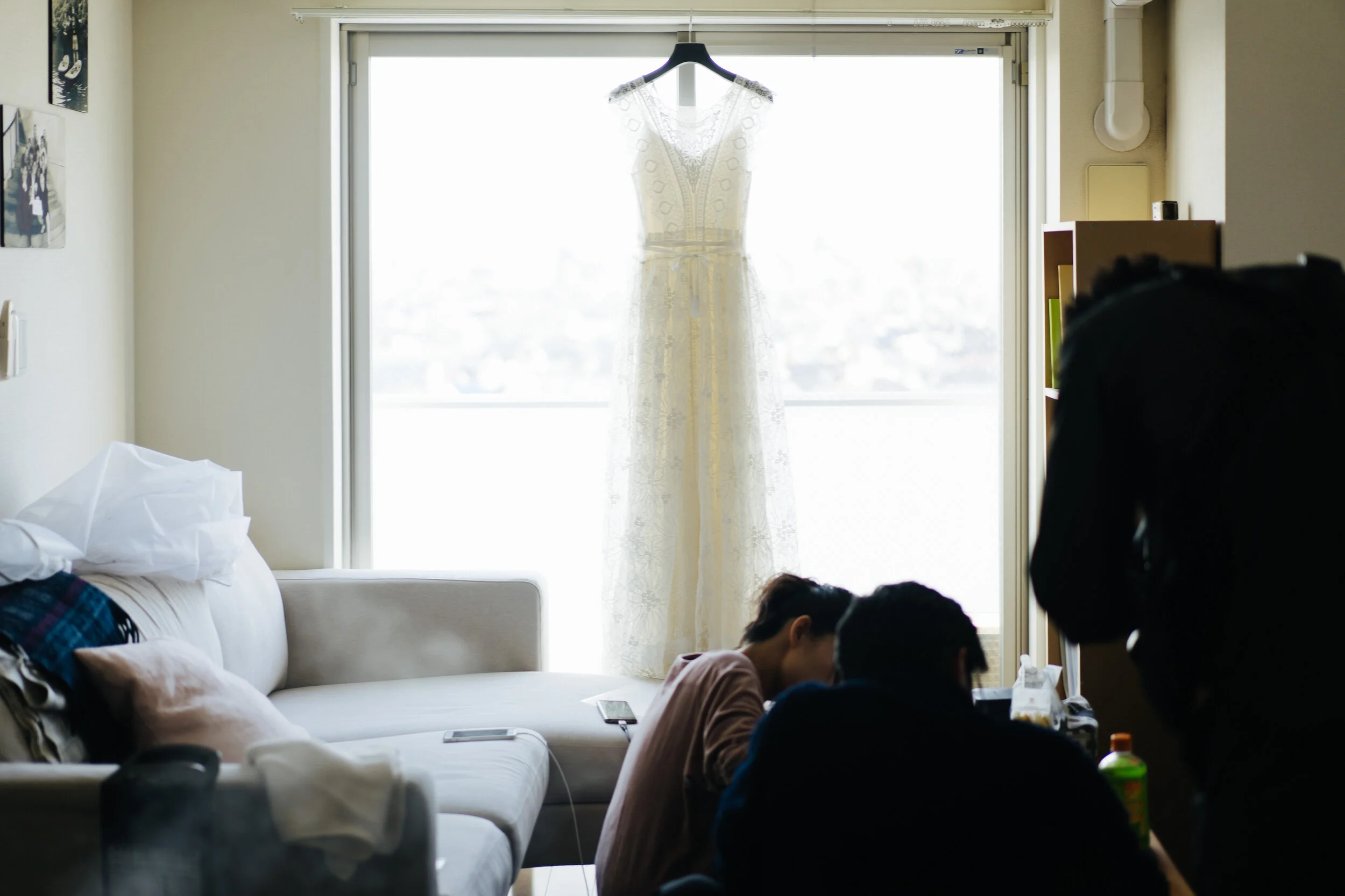 A wedding dress hangs on a black hanger in front of a bright window in a living room, with a white sofa and several people sitting and lying on it, and a bookshelf on the right.