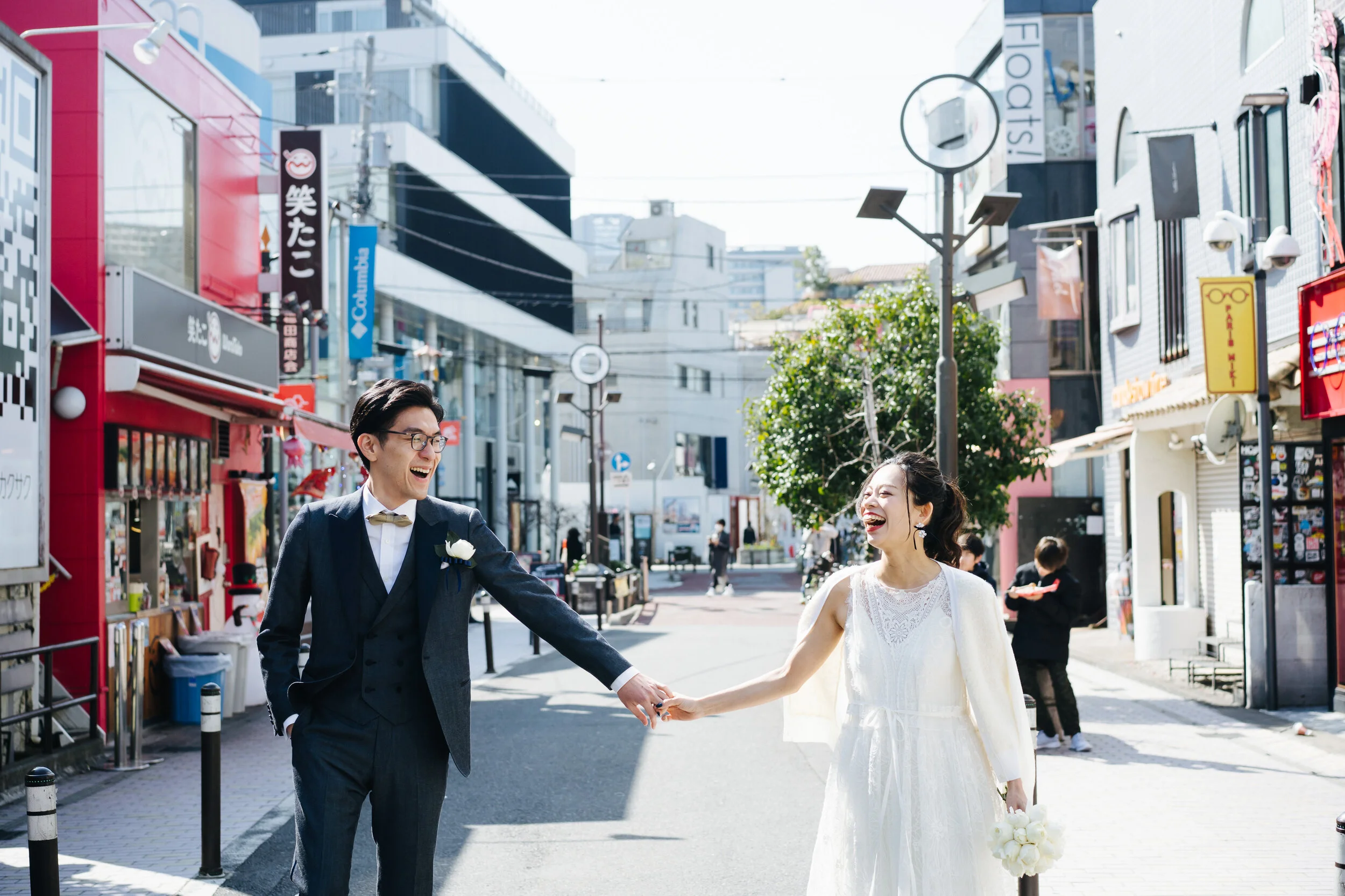 A newlywed couple smiling and holding hands on a city street, with shops and signage in the background.